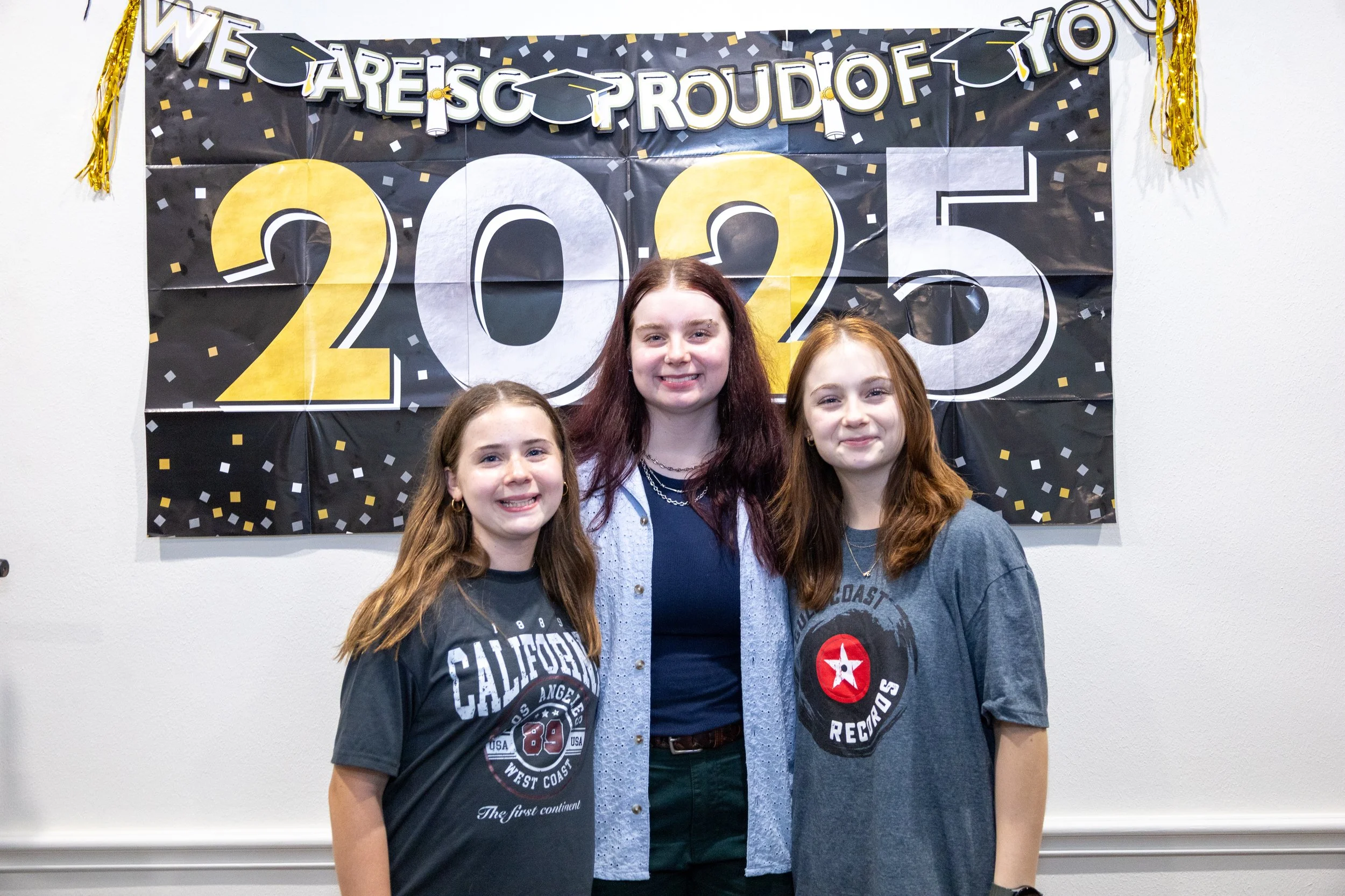 Three young women standing side by side, smiling, in front of a black and yellow graduation celebration banner that reads "WE ARE SO PROUD OF YOU 2025". The woman in the middle has red hair, and the women on either side have light brown hair. They ar
