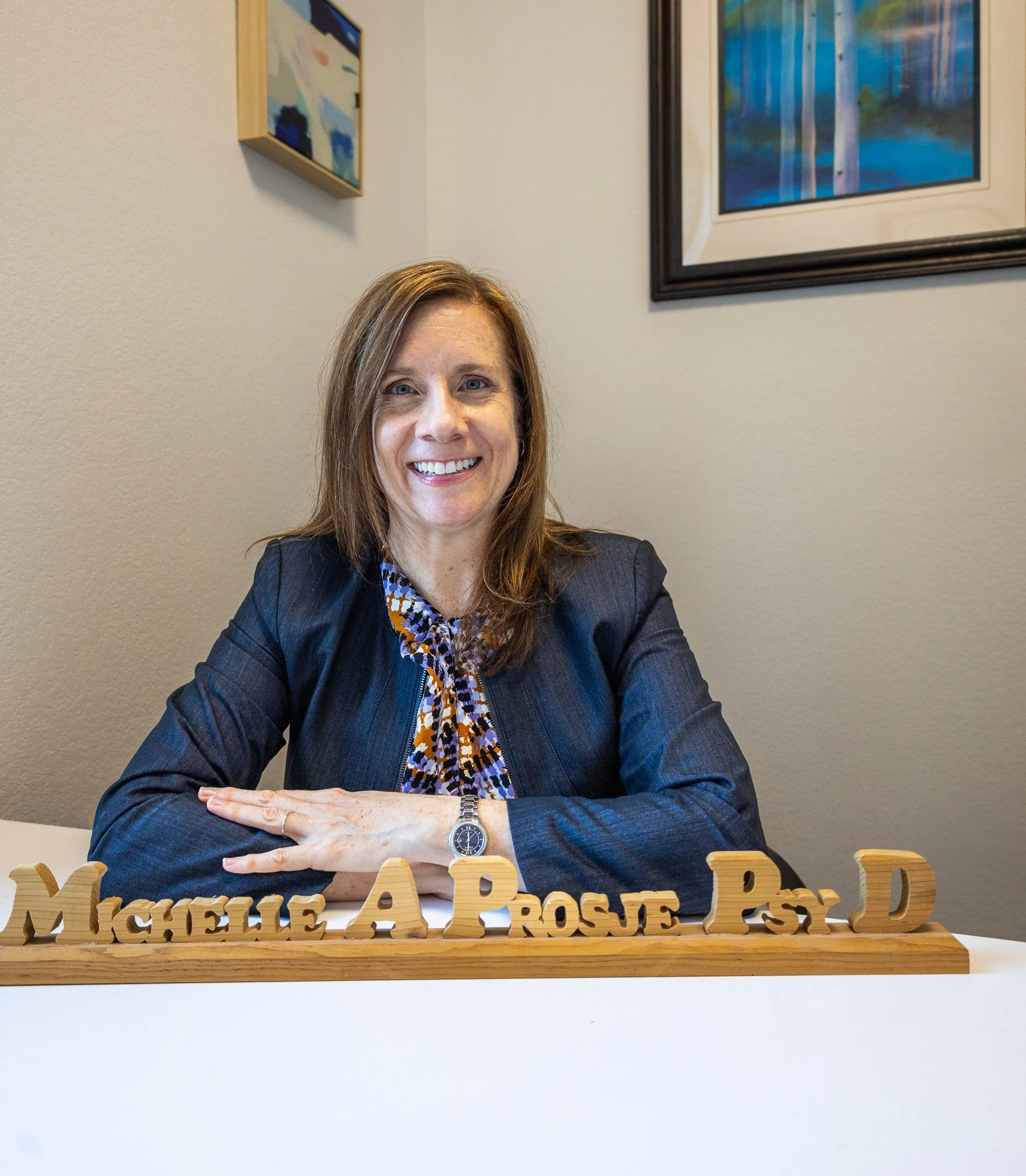 A smiling woman with shoulder-length brown hair sits at a white table with her arms crossed. In front of her are wooden letter blocks spelling out her name and credentials. The background has two framed pieces of artwork on a beige wall.