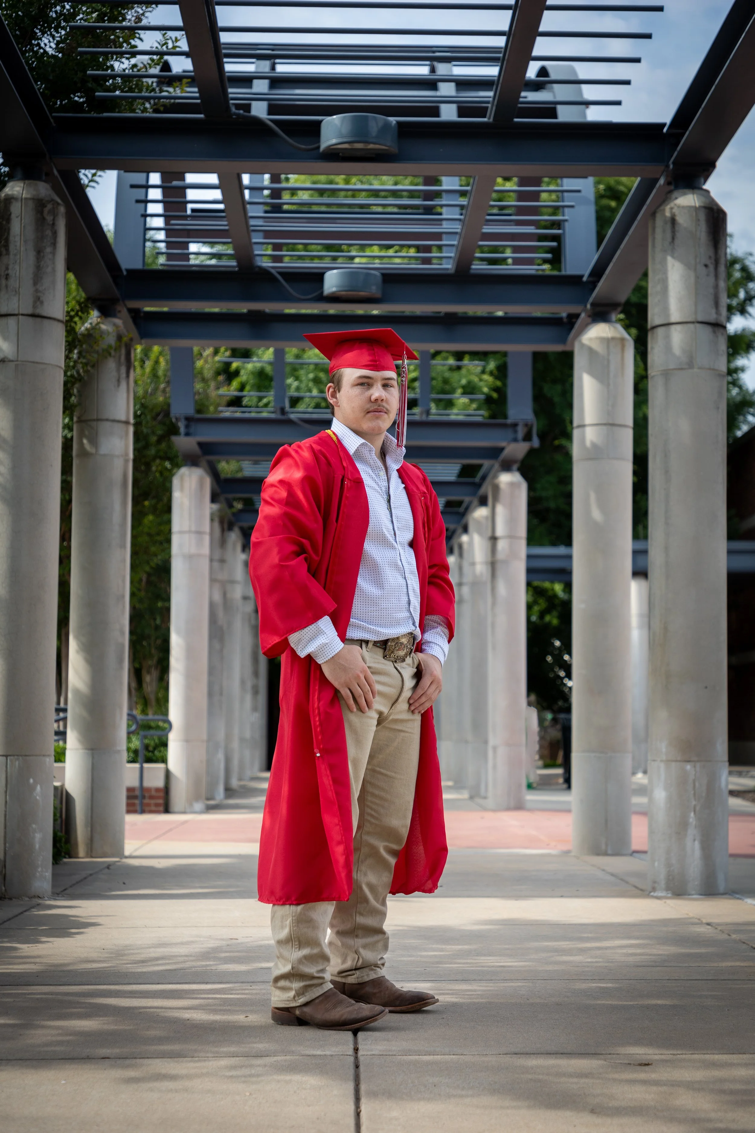 Young man wearing a red graduation cap and gown standing outdoors on a concrete pathway, with concrete pillars and greenery in the background.