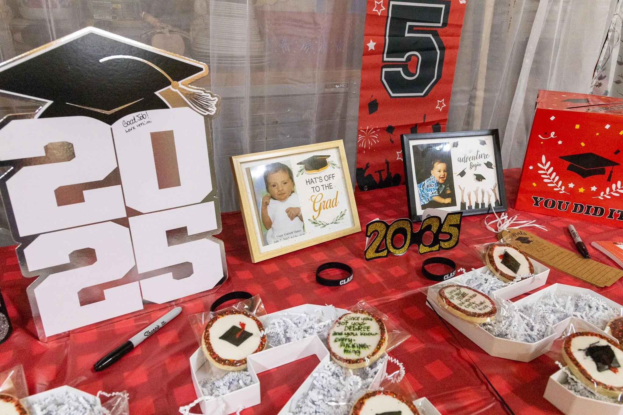 Graduation celebration table with framed photos, cookies, and decorations including a large paper 2023 sign, a red and black graduation-themed banner, and thematic cupcakes.