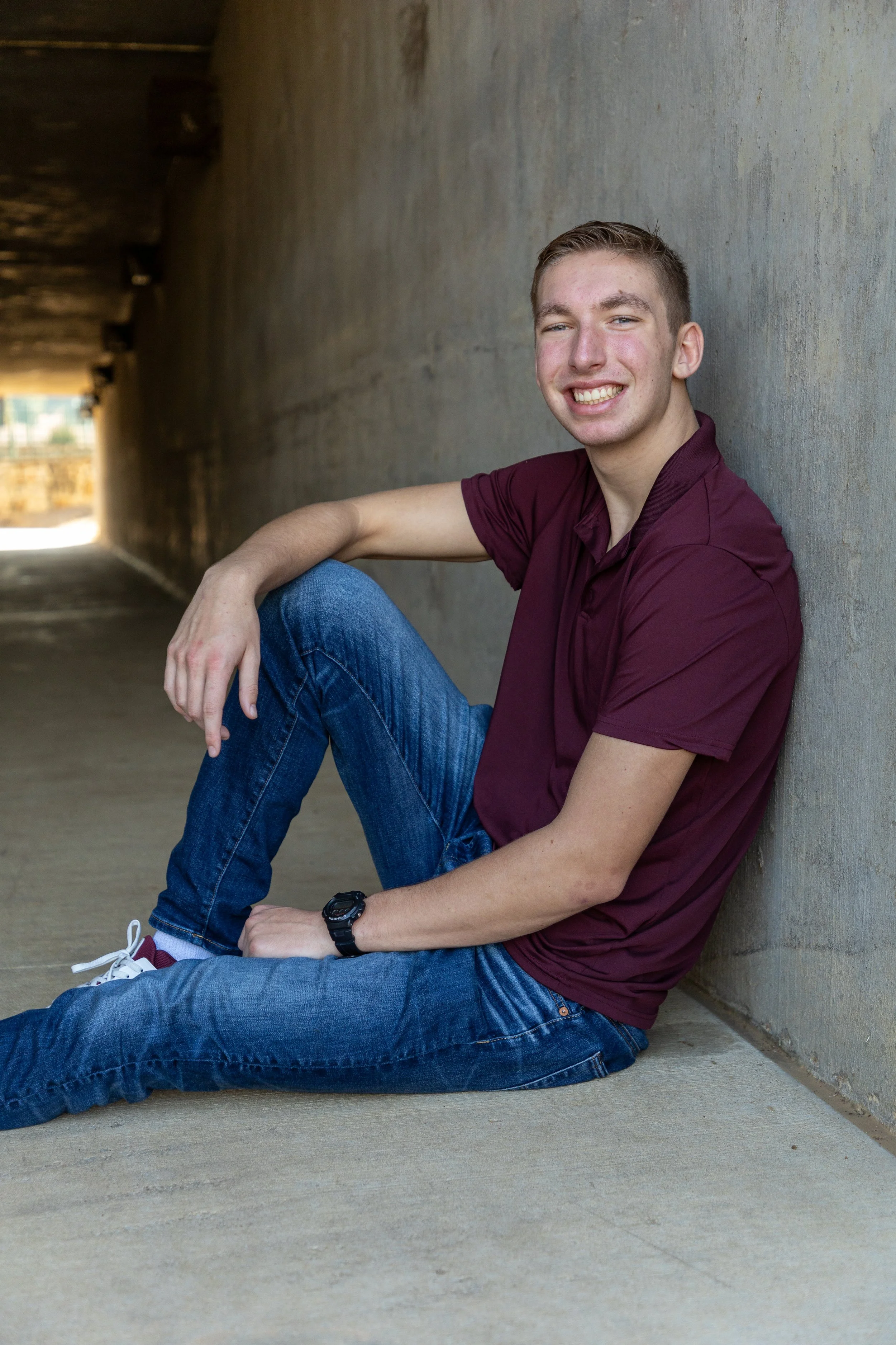 A young man with short light brown hair, smiling and sitting on the ground against a concrete wall in a shaded tunnel, dressed in a maroon polo shirt, blue jeans, white sneakers, and wearing a black watch.