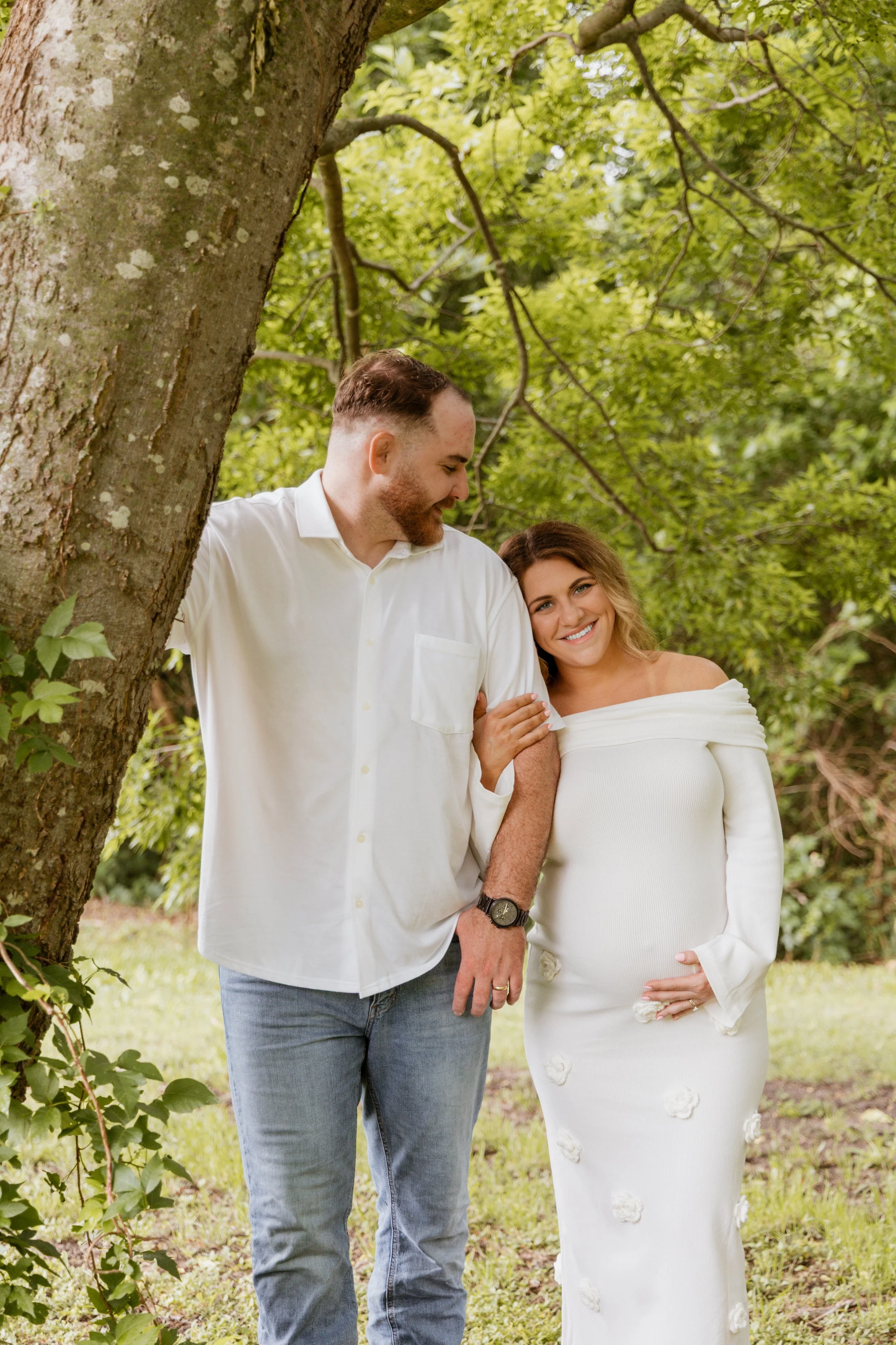 A couple smiling, standing outdoors near a large tree with green leaves, during daytime.