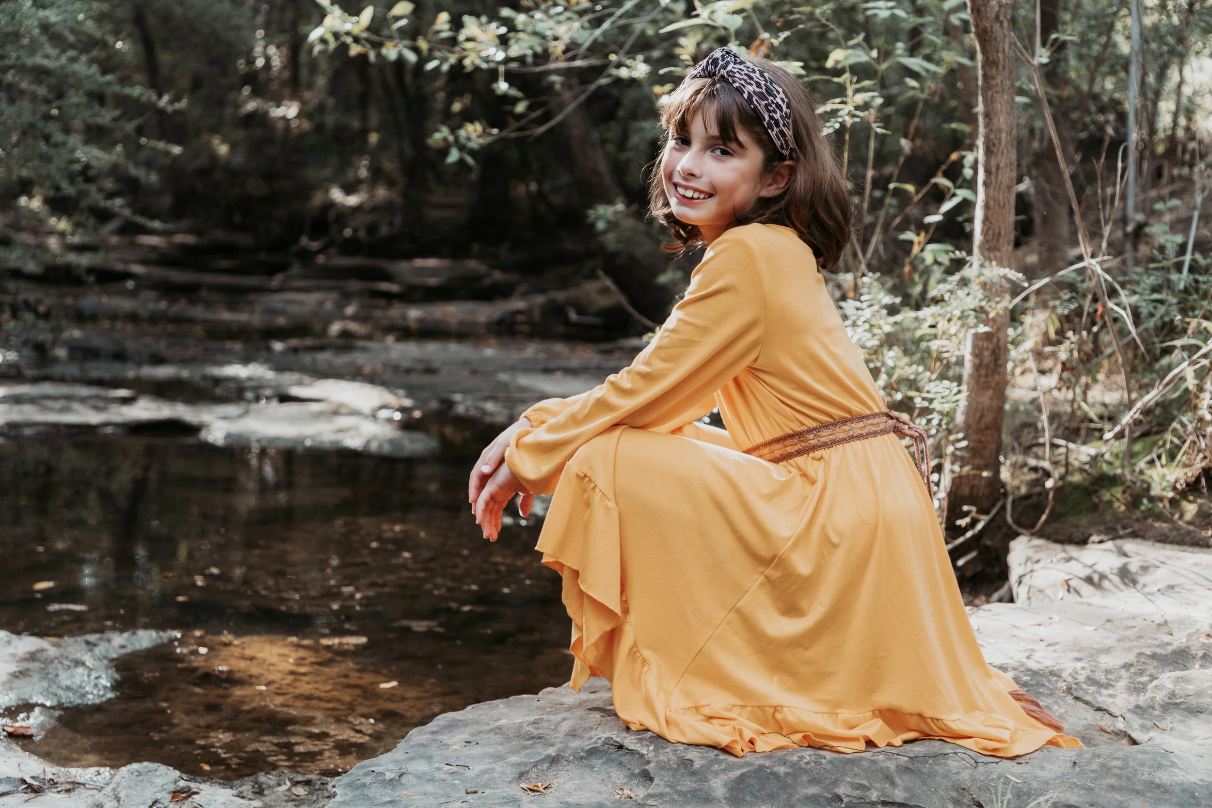 A young girl in a yellow dress sitting on rocks near a stream in a wooded area, smiling and looking at the camera.