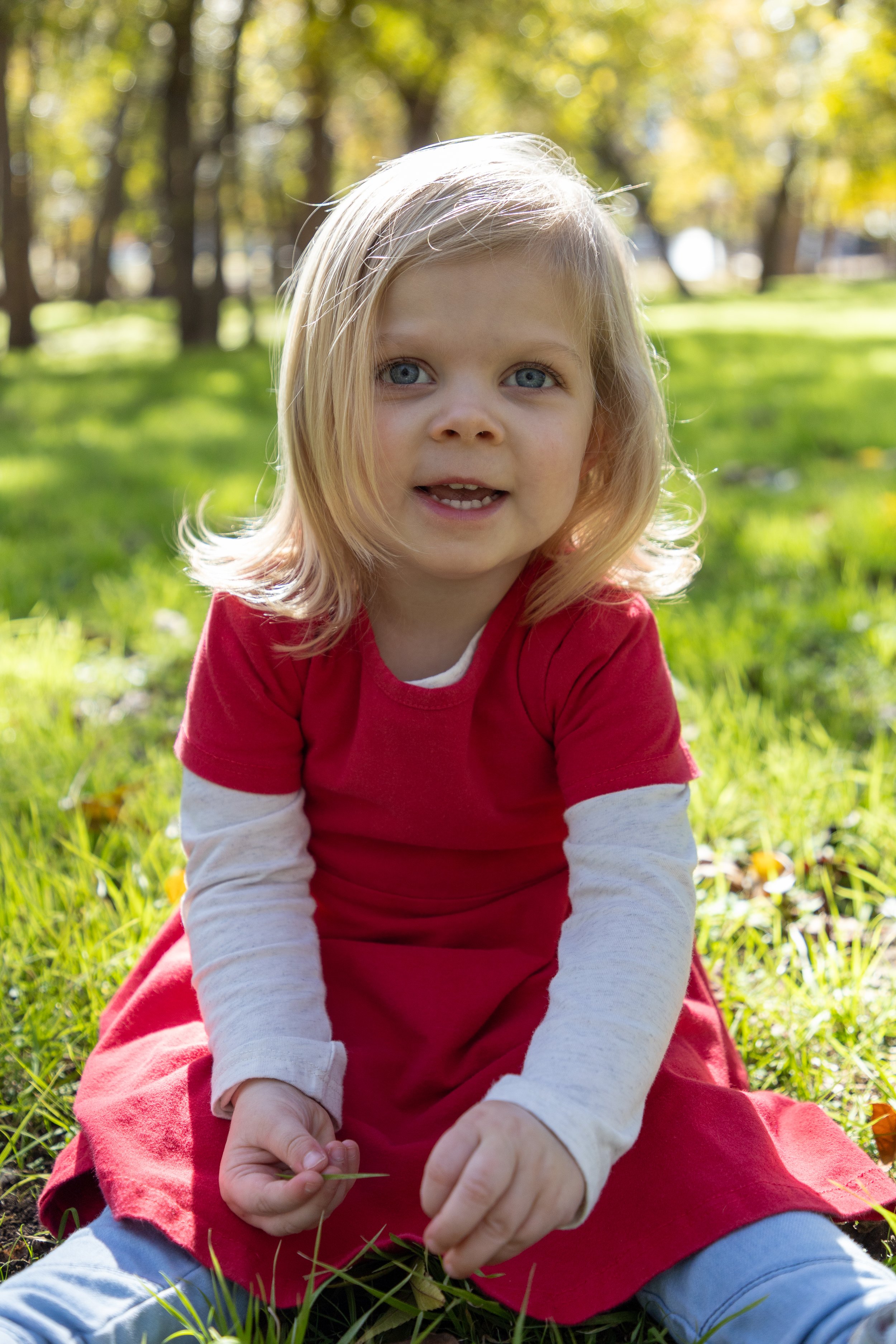A young girl with blonde hair and blue eyes sitting on grass in a park, wearing a red dress and white long-sleeve shirt, holding a small plant, with trees and sunlight in the background.