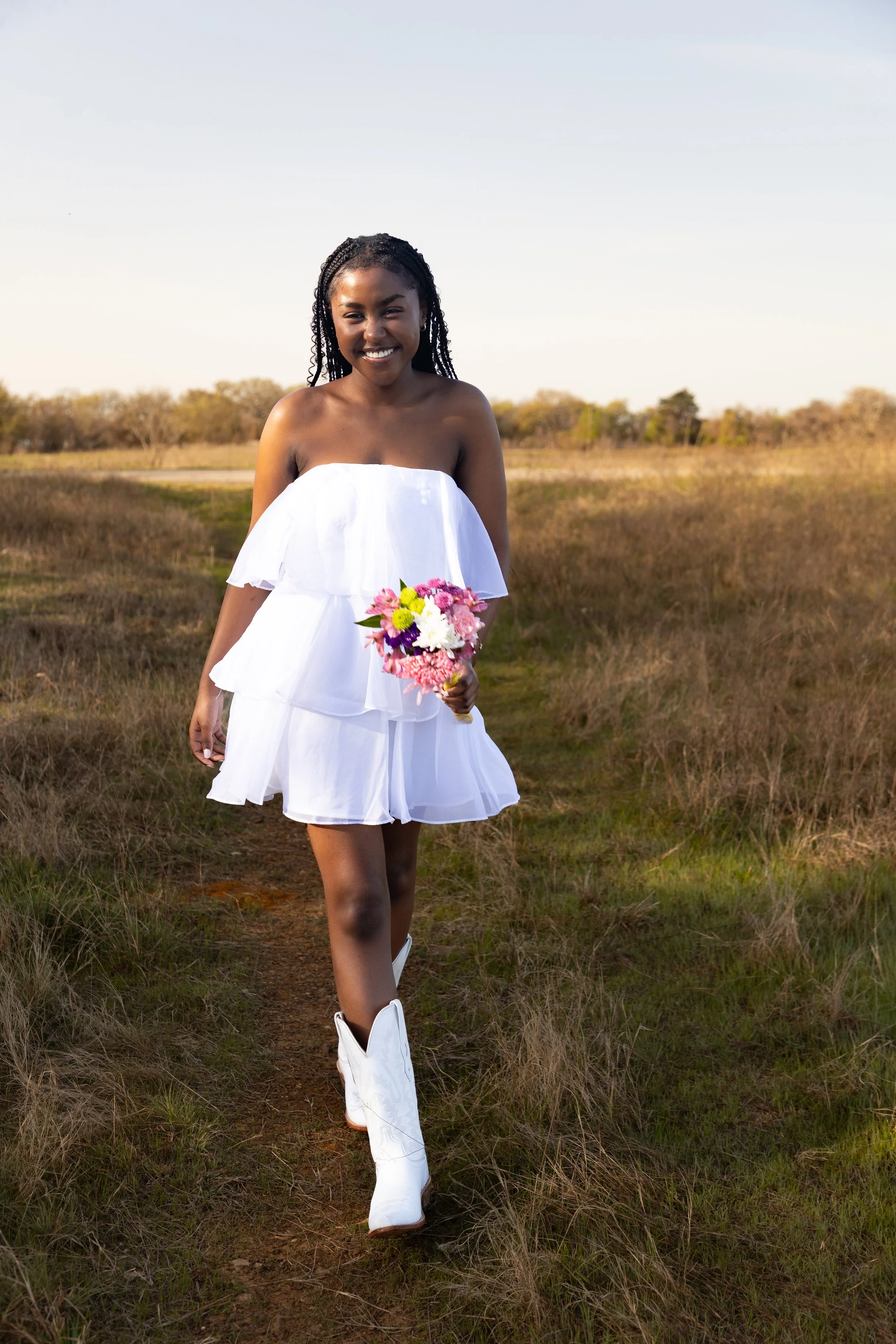Young woman walking on a dirt path in a field, holding a colorful bouquet of flowers, wearing a white strapless dress and white cowboy boots, smiling at the camera.