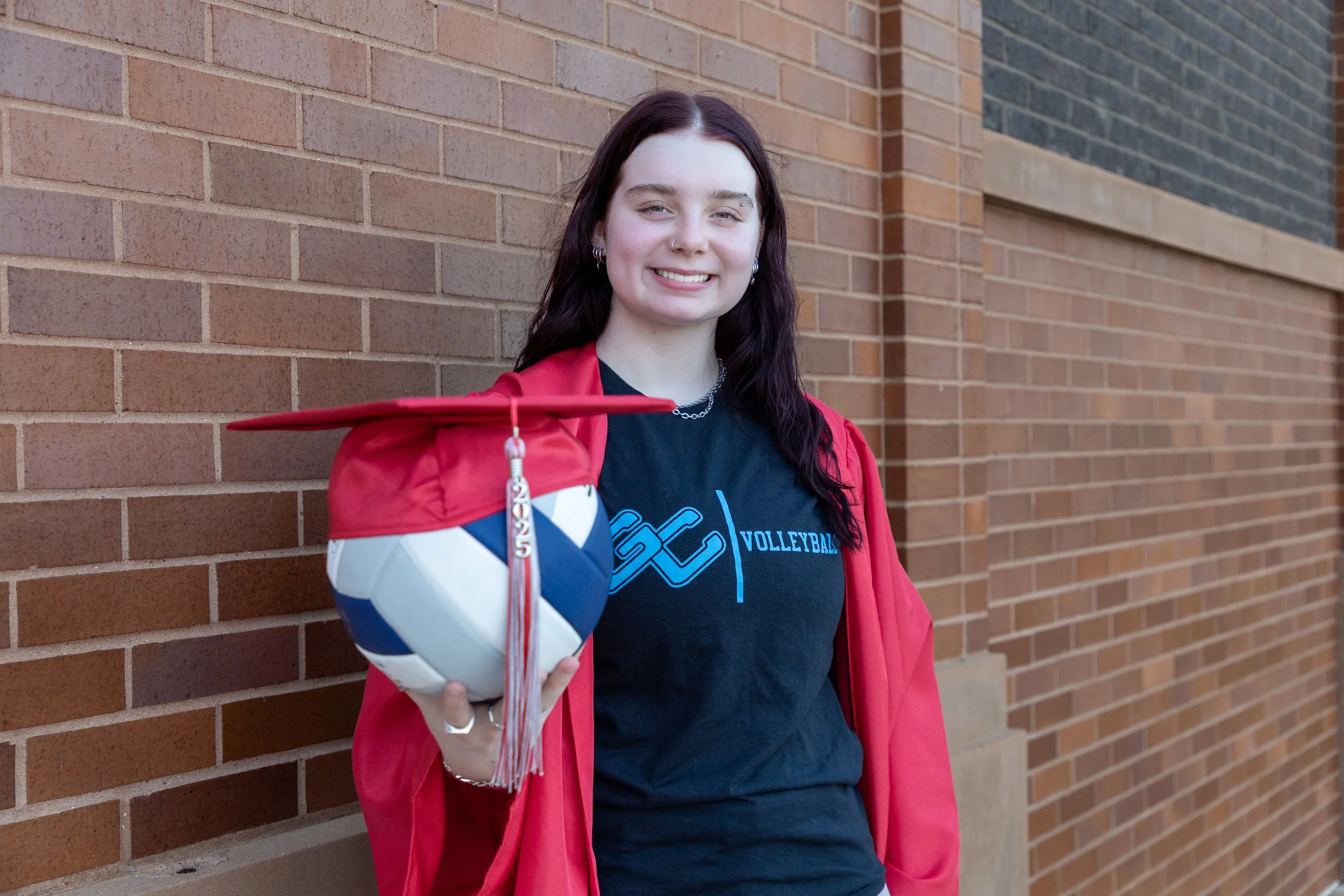 Young woman in a red graduation gown holding a volleyball with her hat resting on her hand, standing outdoors against a brick wall, smiling.