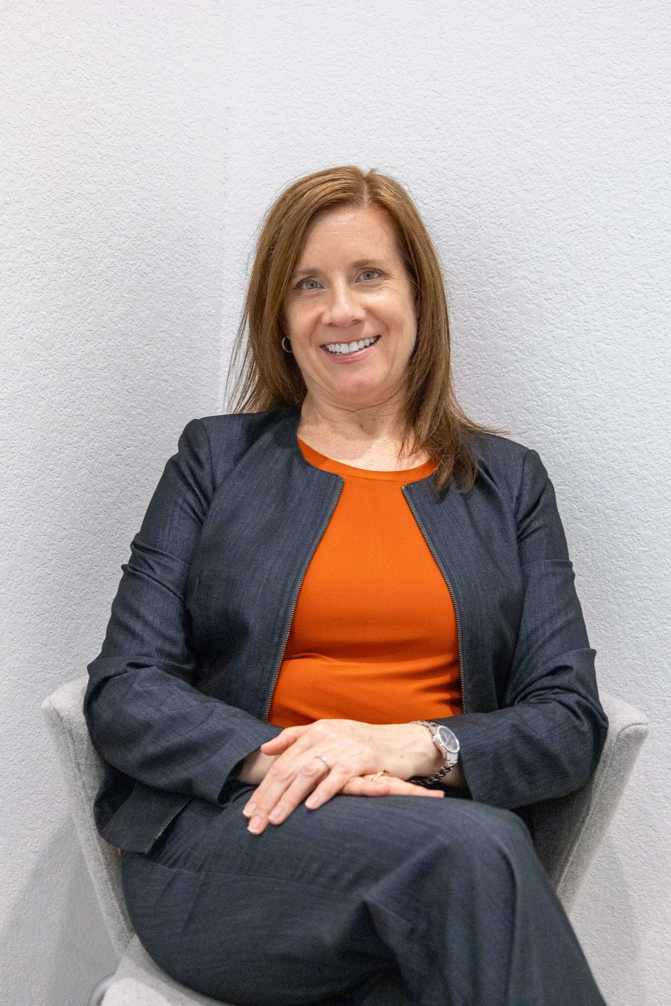 A woman with red hair smiling, sitting in a gray chair against a white textured wall, wearing a black blazer, an orange top, and a watch.
