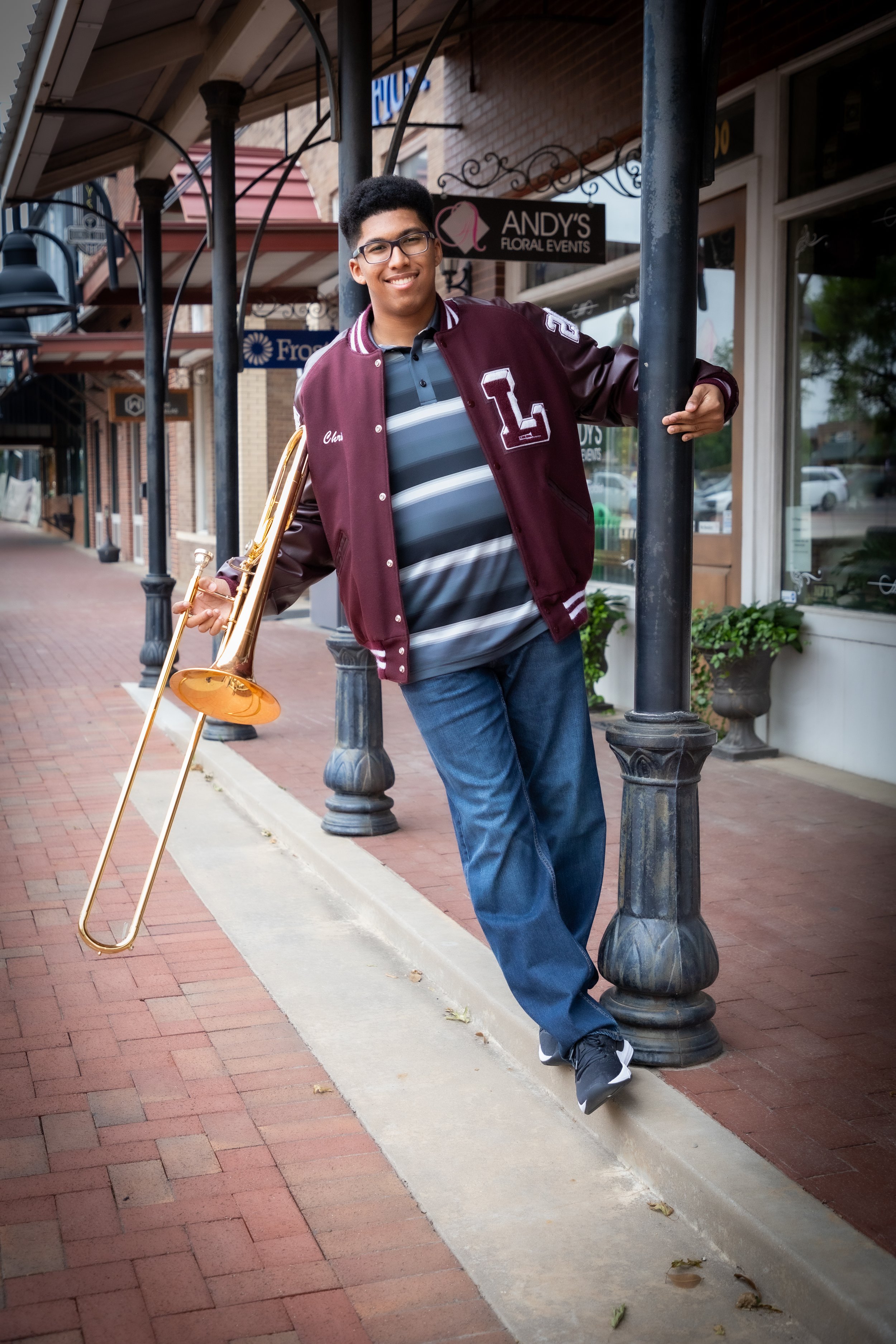 A young man standing on a brick sidewalk holding a trombone, wearing a maroon letterman jacket, striped polo shirt, jeans, sneakers, and glasses, smiling at the camera.