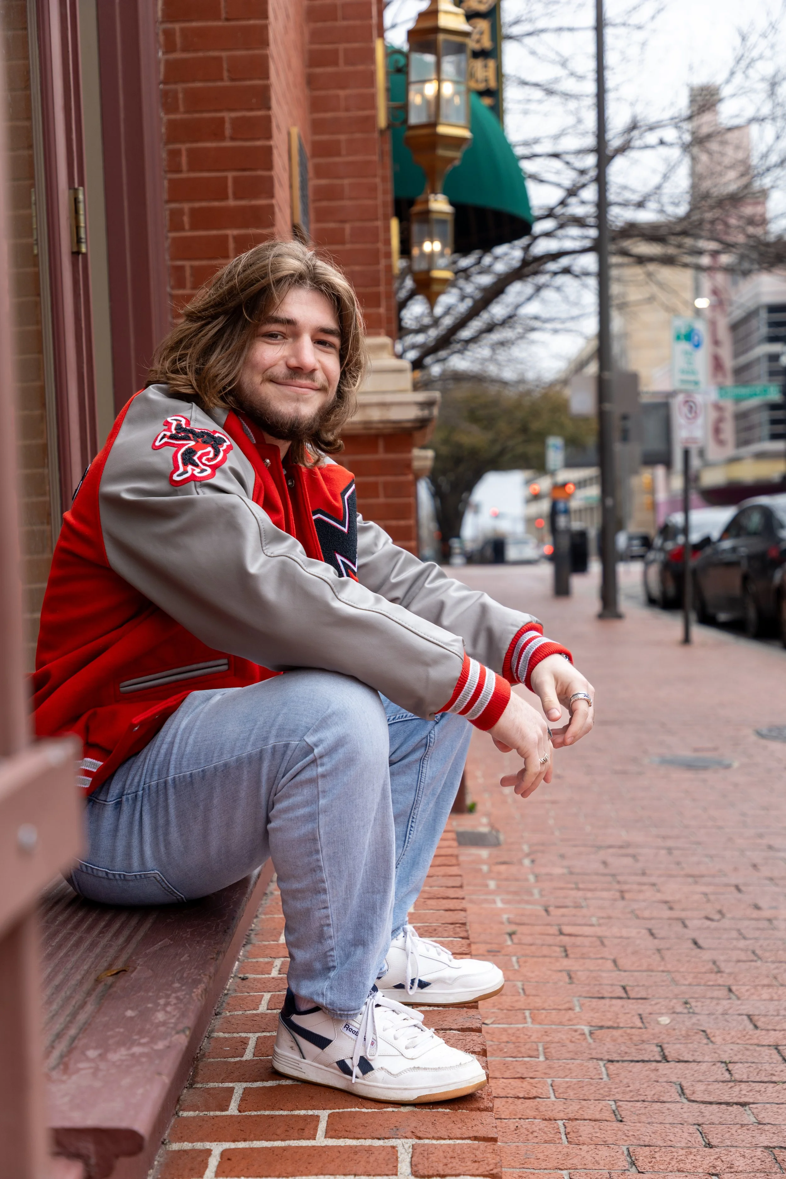 A young man with shoulder-length hair and a beard sitting on a brick sidewalk outside a building, wearing a red and gray varsity jacket, light blue jeans, and white sneakers, smiling at the camera.