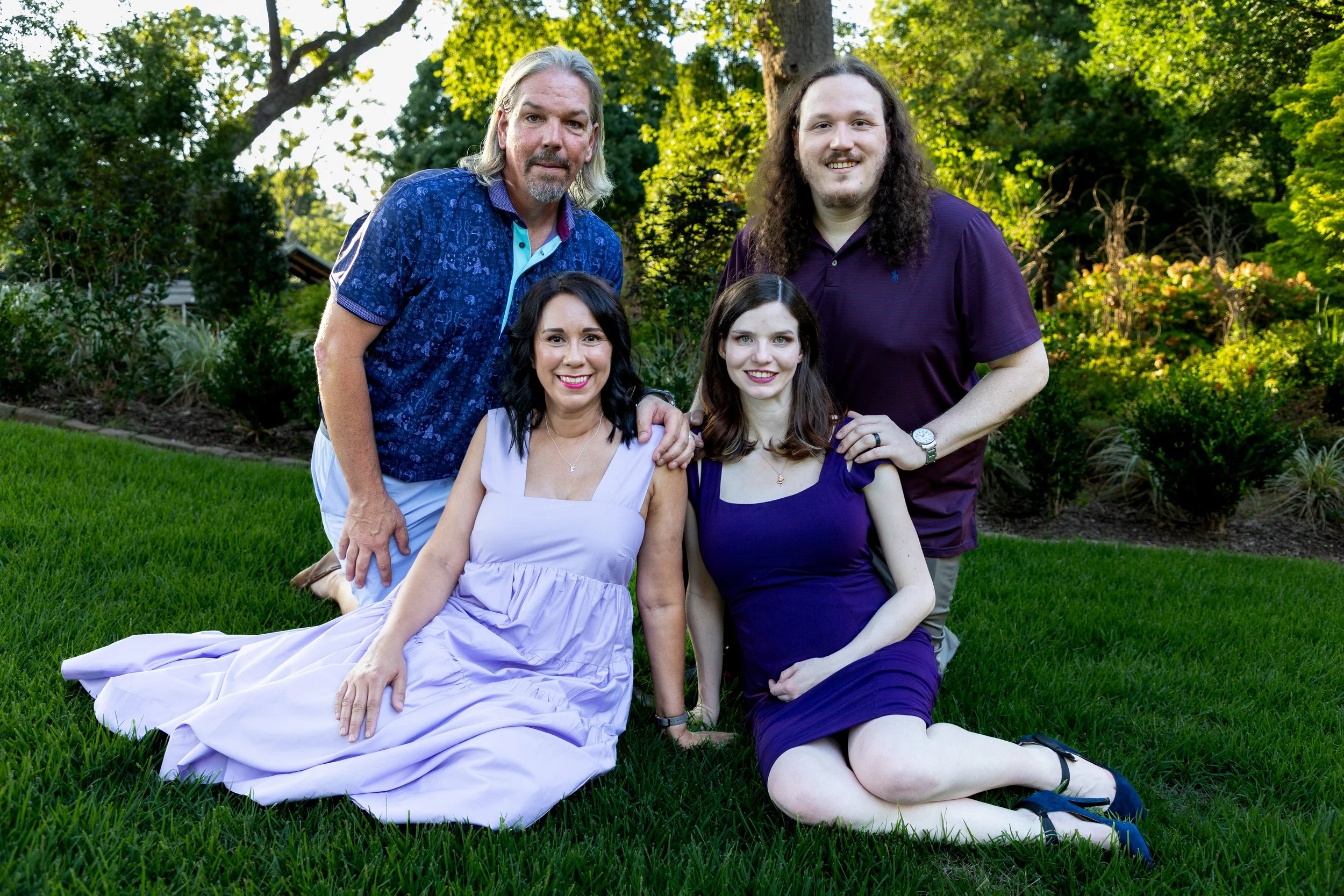 A group of four people outdoors on a lawn with greenery in the background, two women sitting on the grass and two men standing behind them, smiling at the camera.