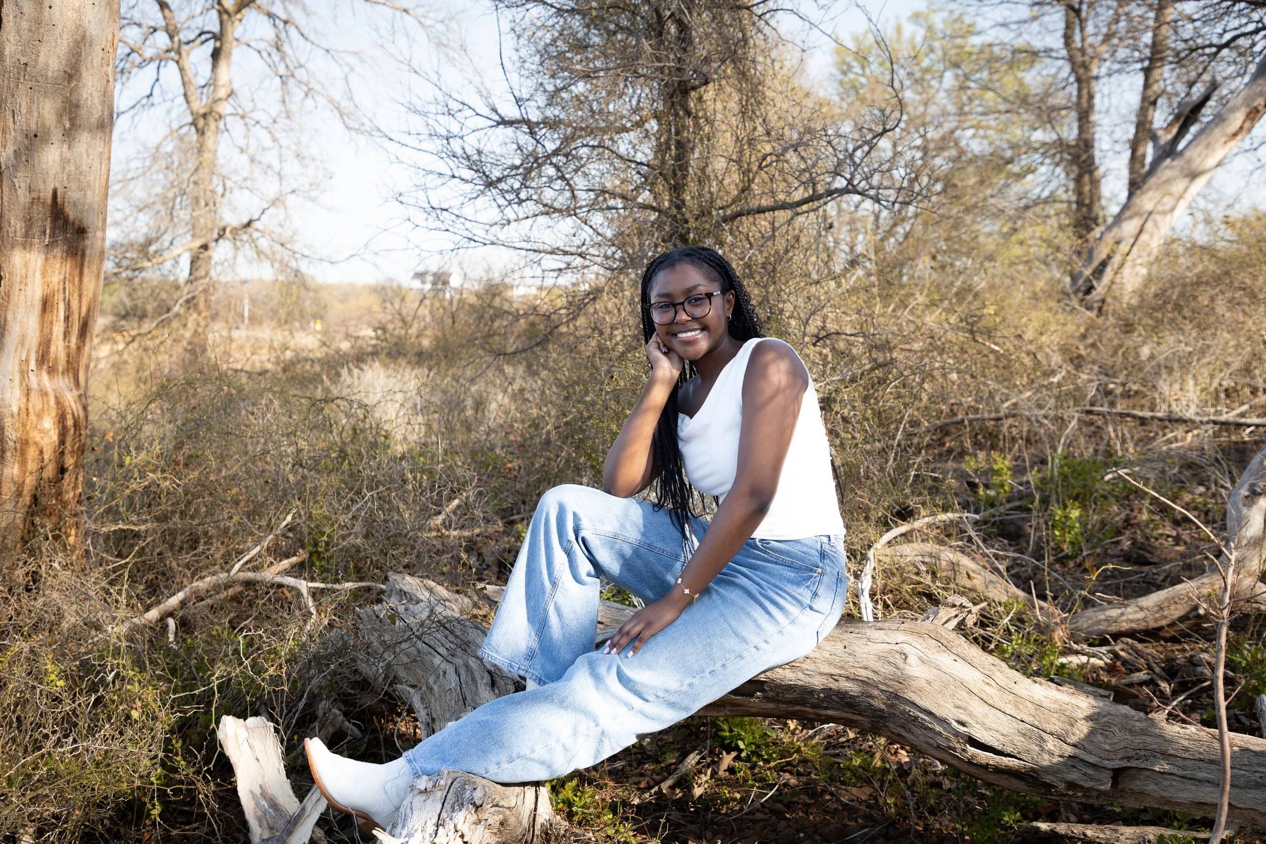 A young woman with glasses, wearing a white sleeveless top and light blue jeans, sitting on a fallen tree trunk in a natural outdoor setting with trees and bushes in the background, smiling and resting her chin on her hand.