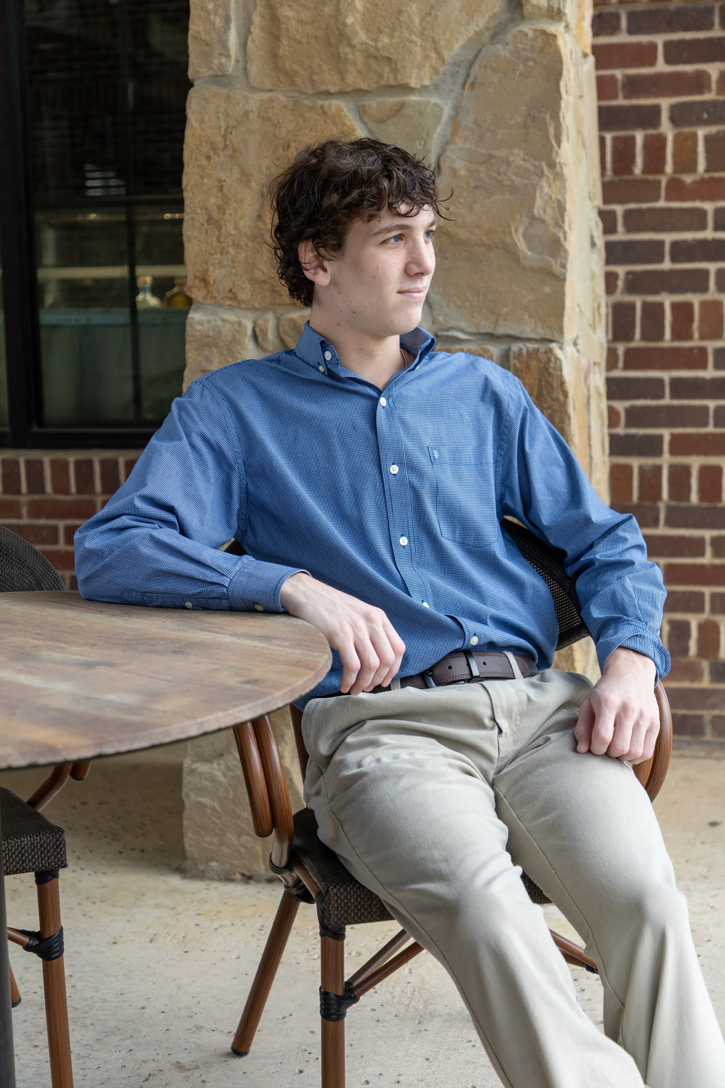 A young man with curly brown hair, wearing a blue button-up shirt and beige pants, sitting at an outdoor table, looking to the right with a thoughtful expression.