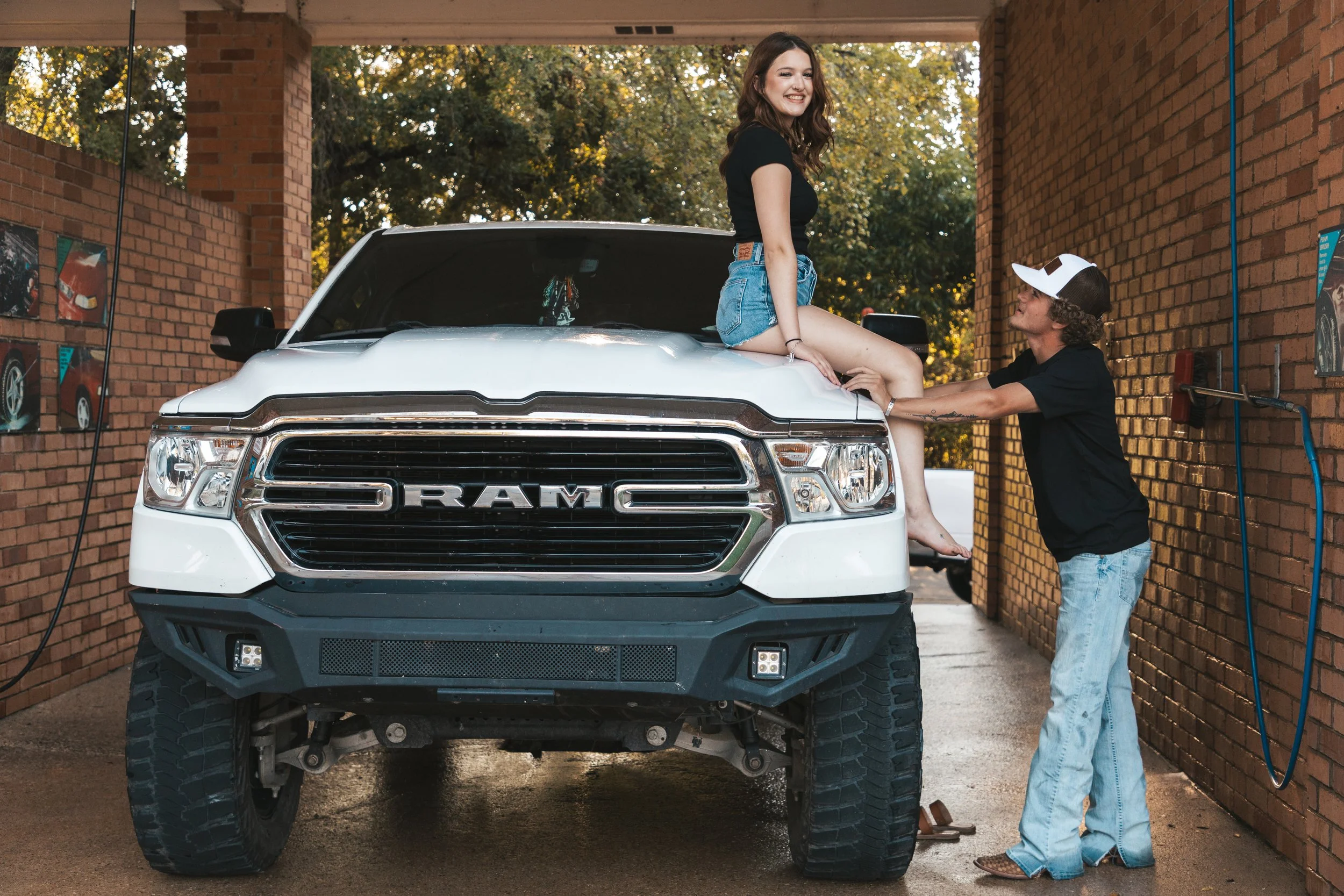 A young woman sitting on the hood of a white RAM truck at a car wash, smiling, while a man in a black t-shirt and blue jeans cleans her feet.