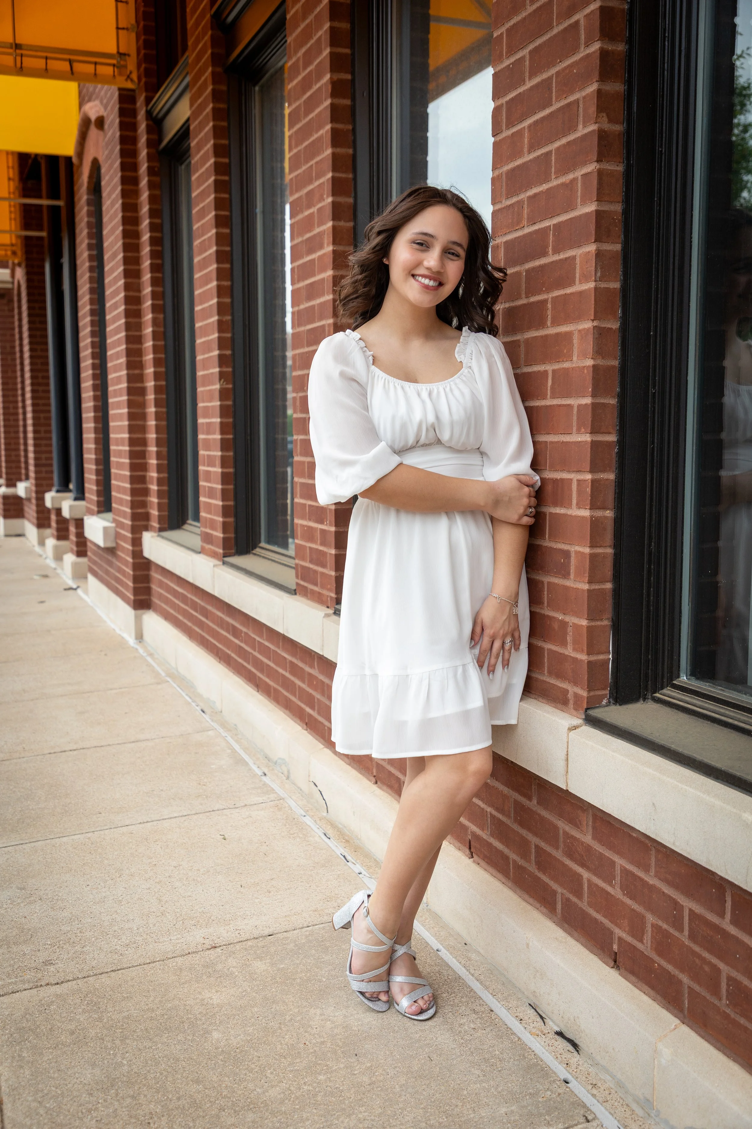 A young woman in a white dress and silver heels standing against a brick building, smiling at the camera.