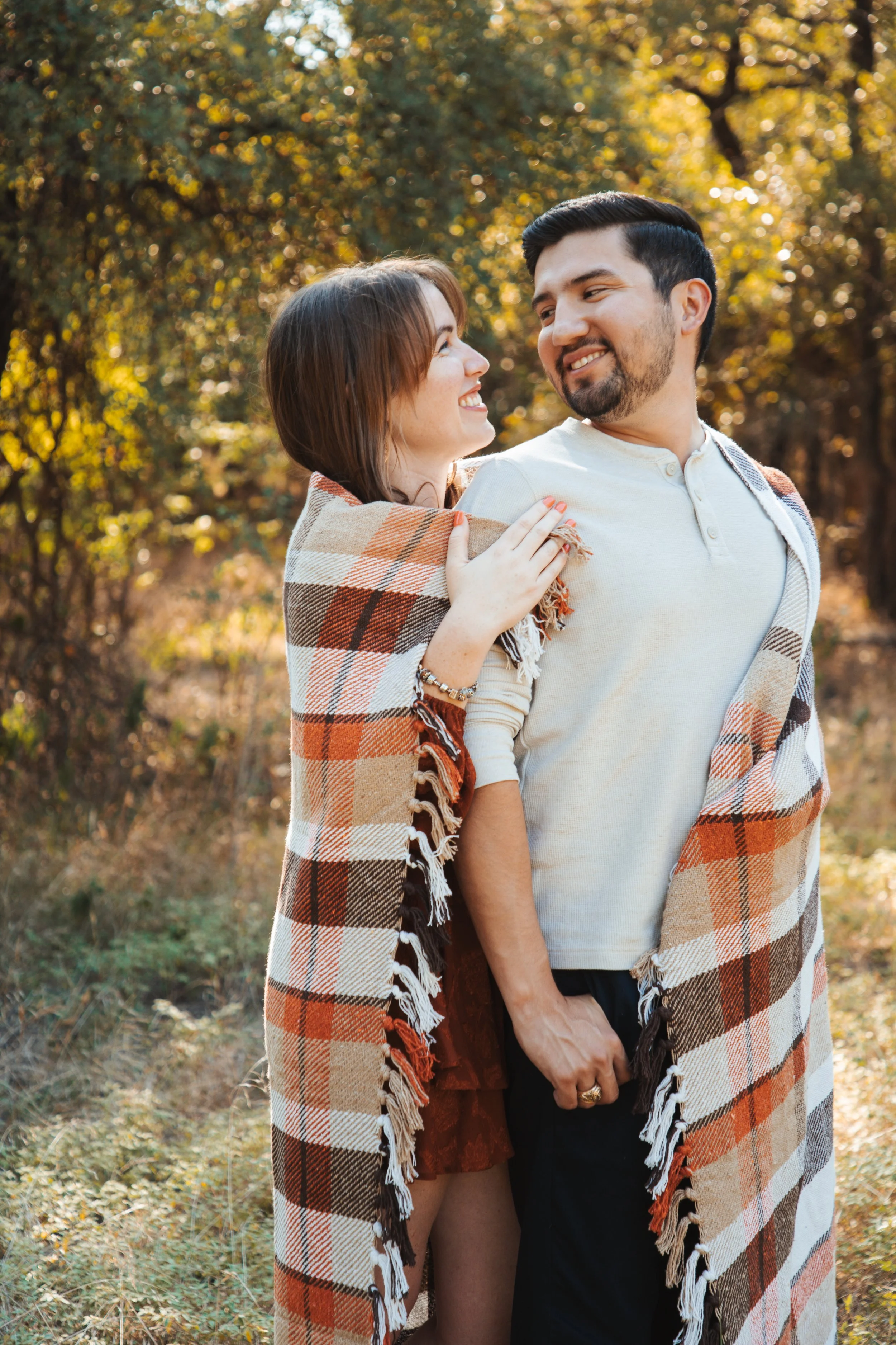 A couple smiling warmly at each other outdoors, wrapped in blankets, with trees and autumn foliage in the background.