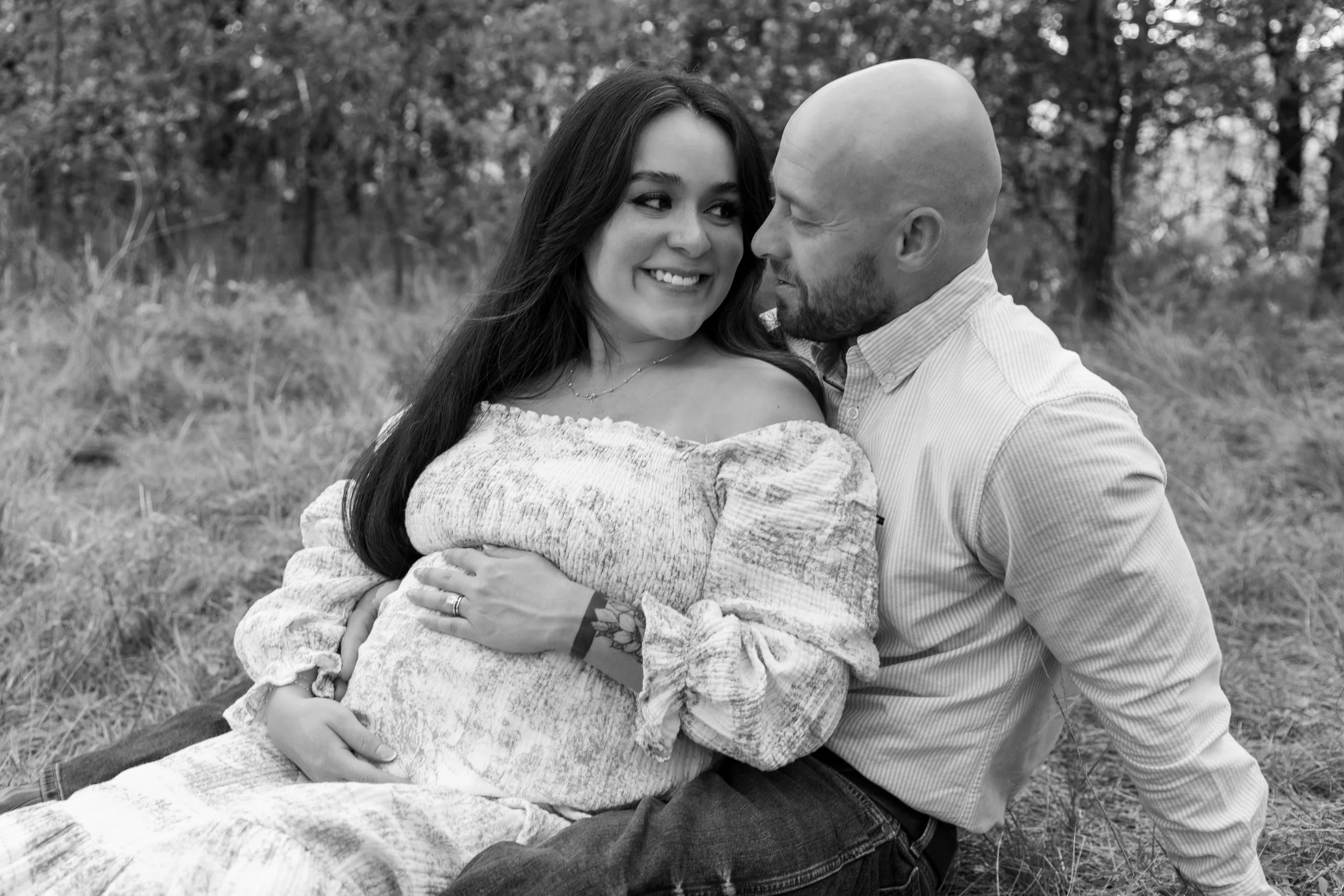 A couple sitting outdoors with trees in the background, the woman is pregnant and smiling at the man, who is looking at her affectionately, in a black and white photo.