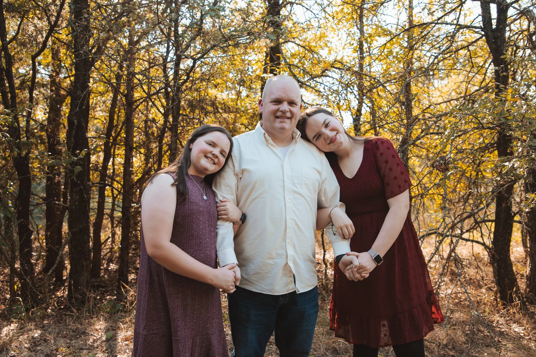 A family of three standing close together outdoors during autumn, surrounded by trees with yellow and brown leaves. The father is in the middle, wearing a cream-colored shirt, with two daughters on either side. The daughter on the left is wearing a p