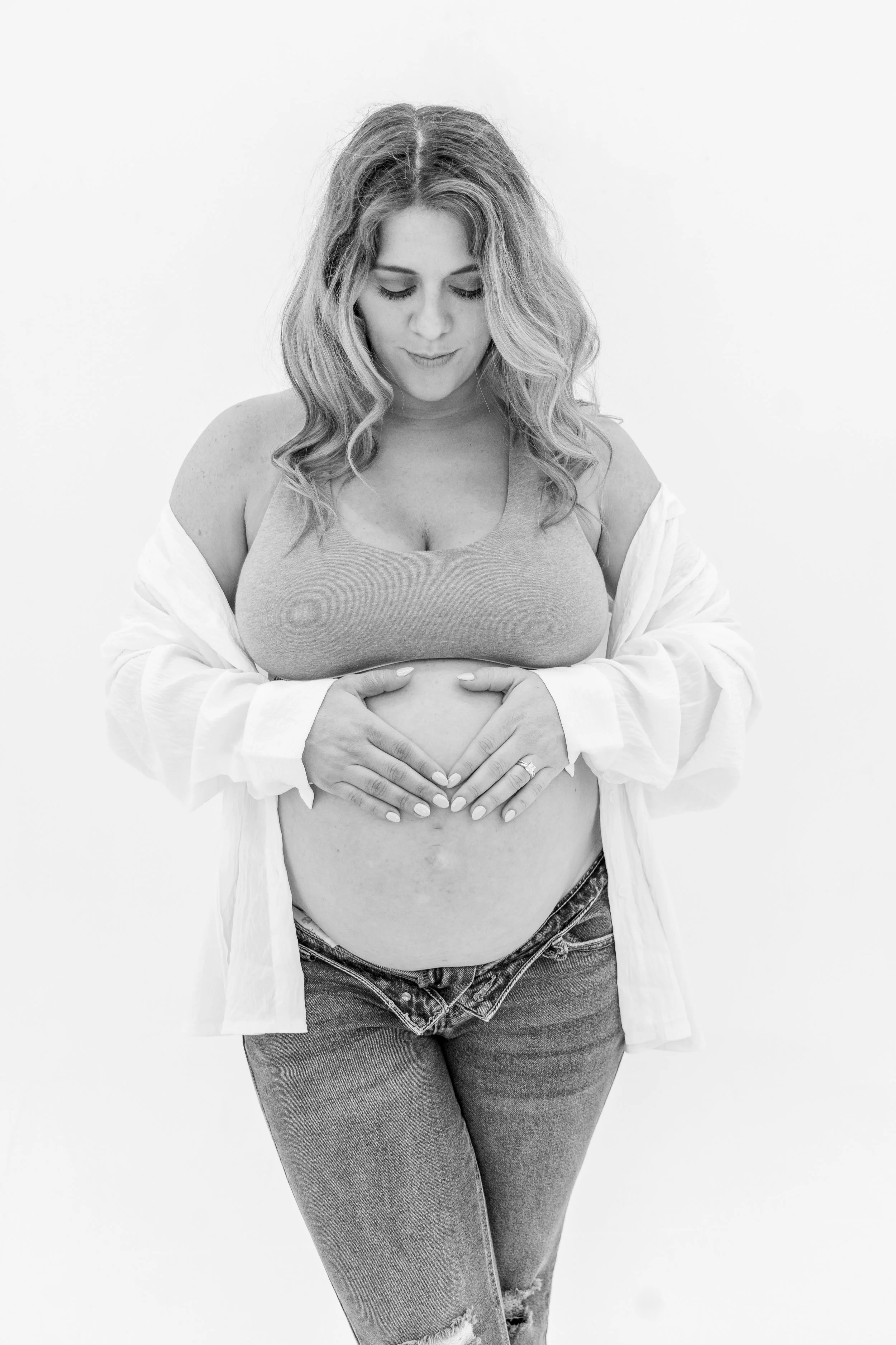 Black and white photograph of a pregnant woman with wavy hair, wearing a tank top and jeans, holding her belly with both hands, and standing against a plain white background.