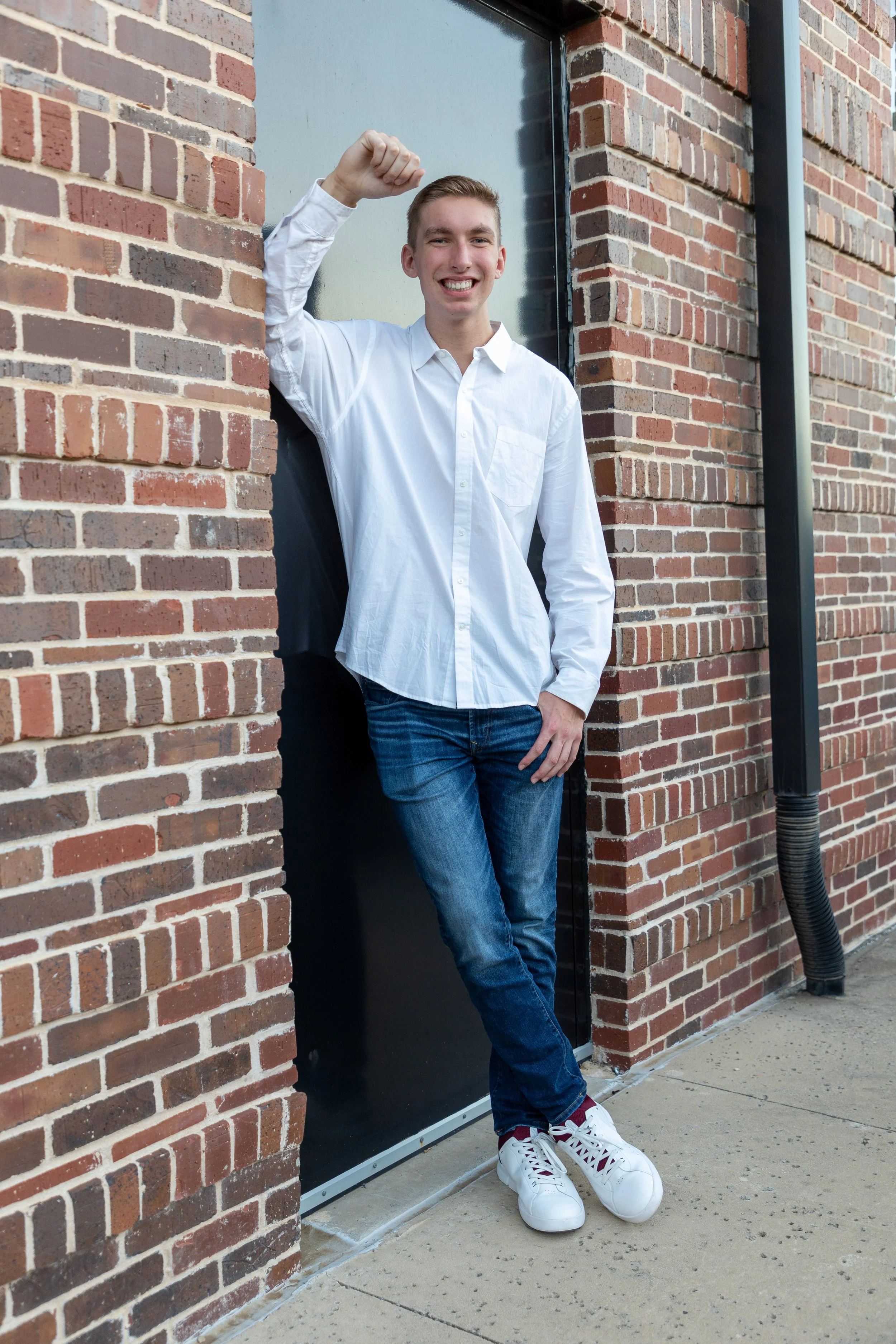 A young man with short blonde hair, wearing a white button-up shirt, blue jeans, and white sneakers, smiling and raising his right fist, standing outdoors against a brick wall next to a black door.