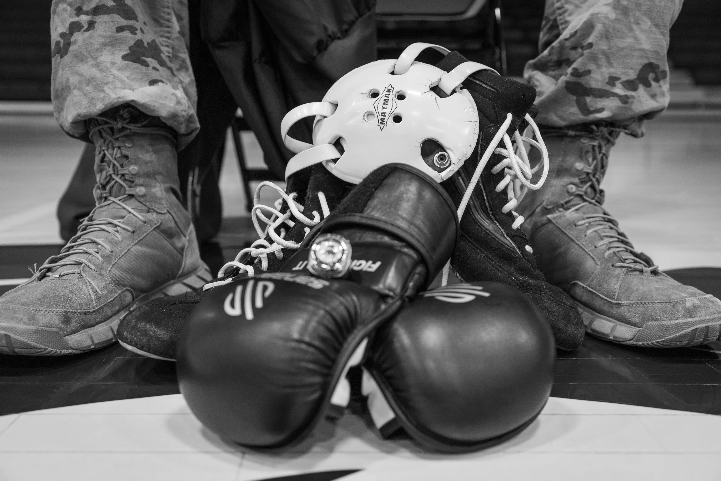 Boxing gloves, a protective headgear, and a pair of tactical boots on a boxing ring floor. The items are black, white, and grey, with the headgear and gloves showing signs of wear.