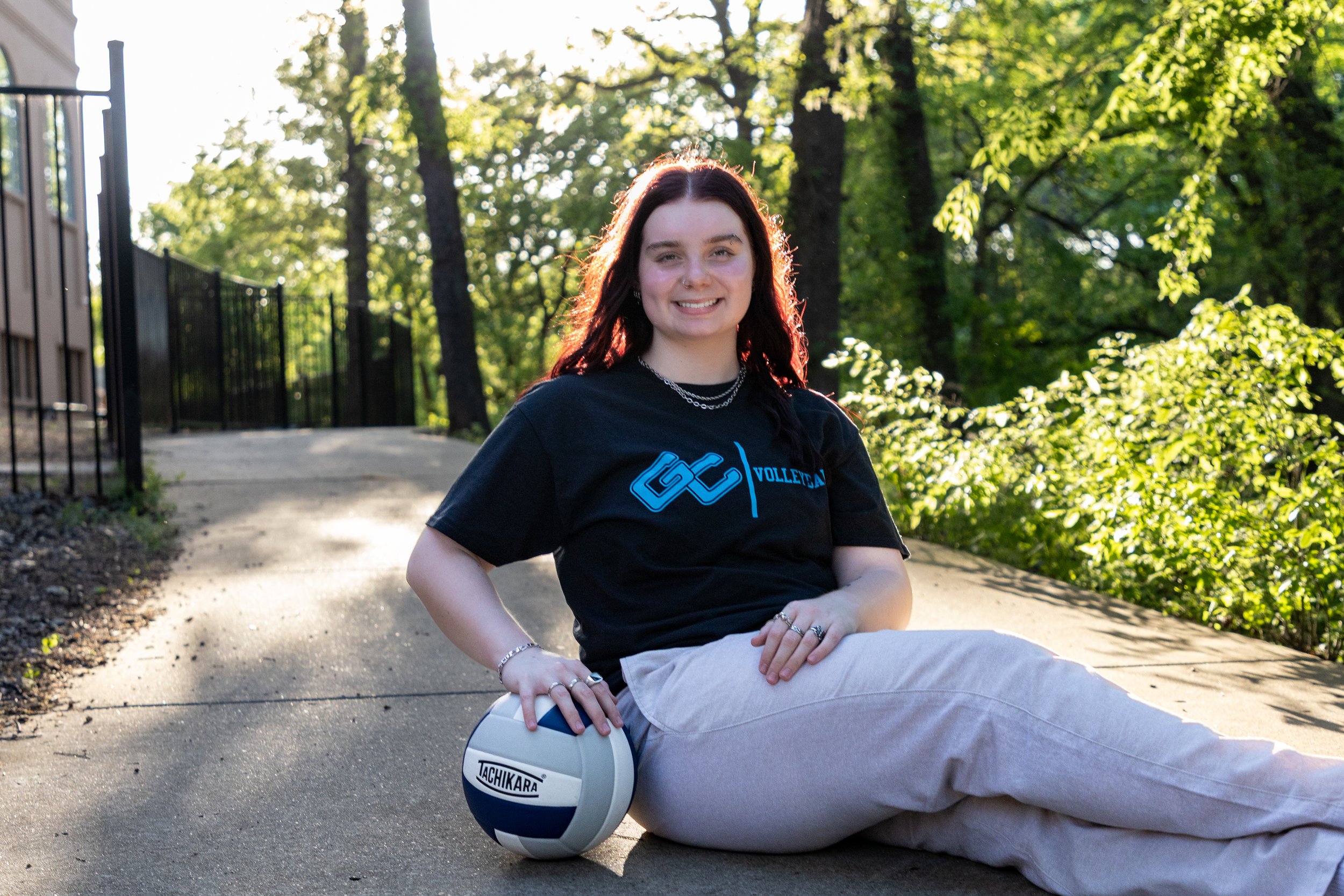 A young woman with red hair and jewelry, smiling while sitting on the sidewalk outdoors in a wooded area, with her arm resting on a volleyball labeled Tachikara.