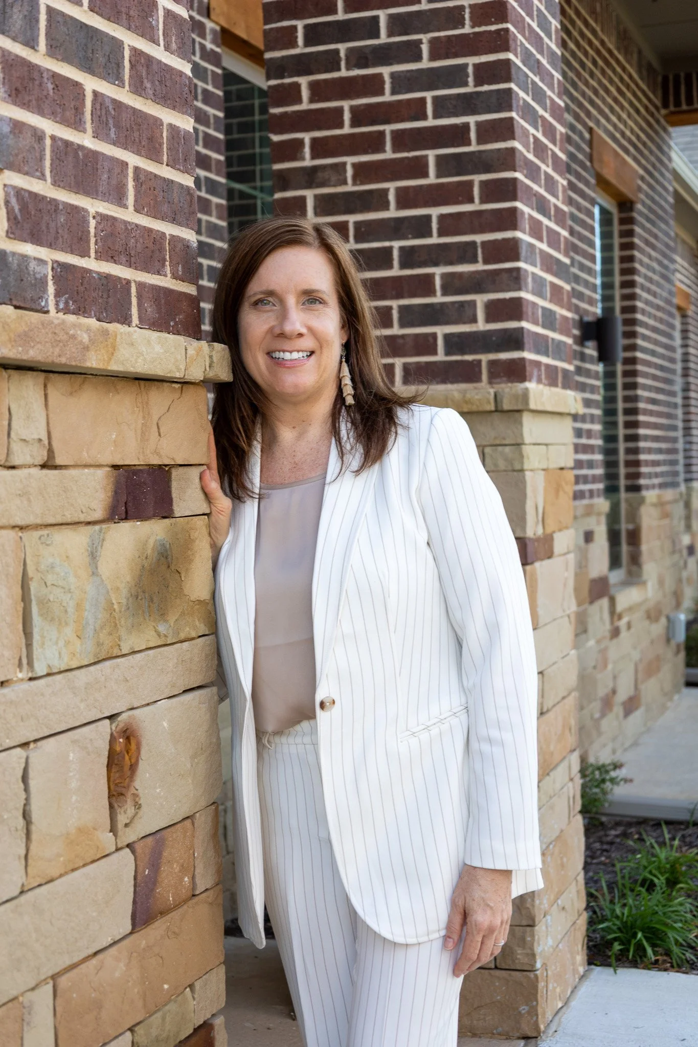 A woman in a white pinstripe suit smiling and leaning against a stone and brick wall outside a building.