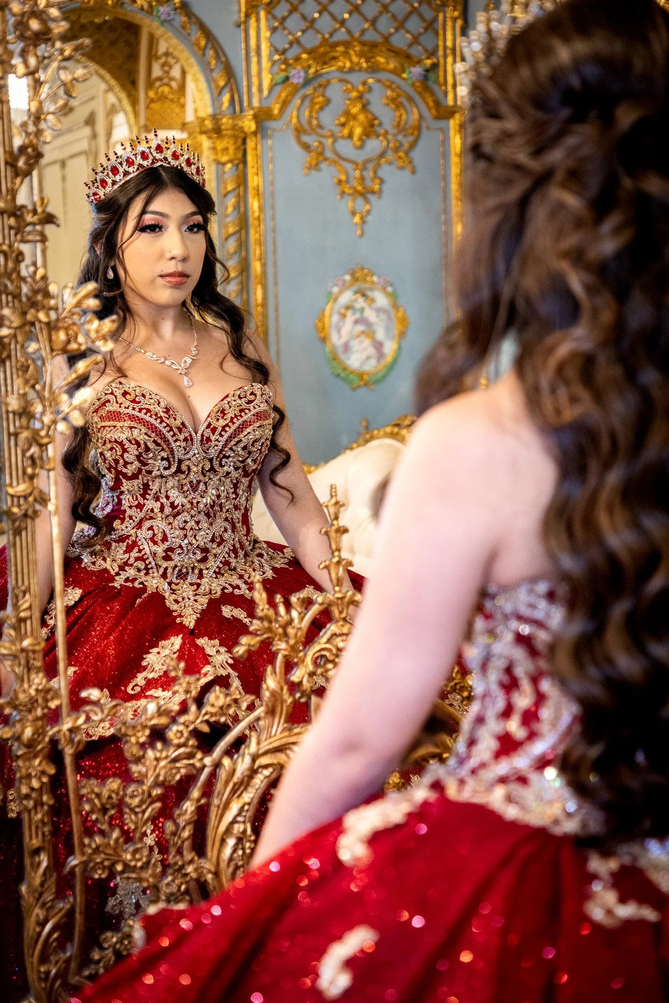 A woman in an ornate red and gold dress wearing a crown, looking at herself in a mirror with a gold frame, in a luxurious room with detailed gold and blue decor.