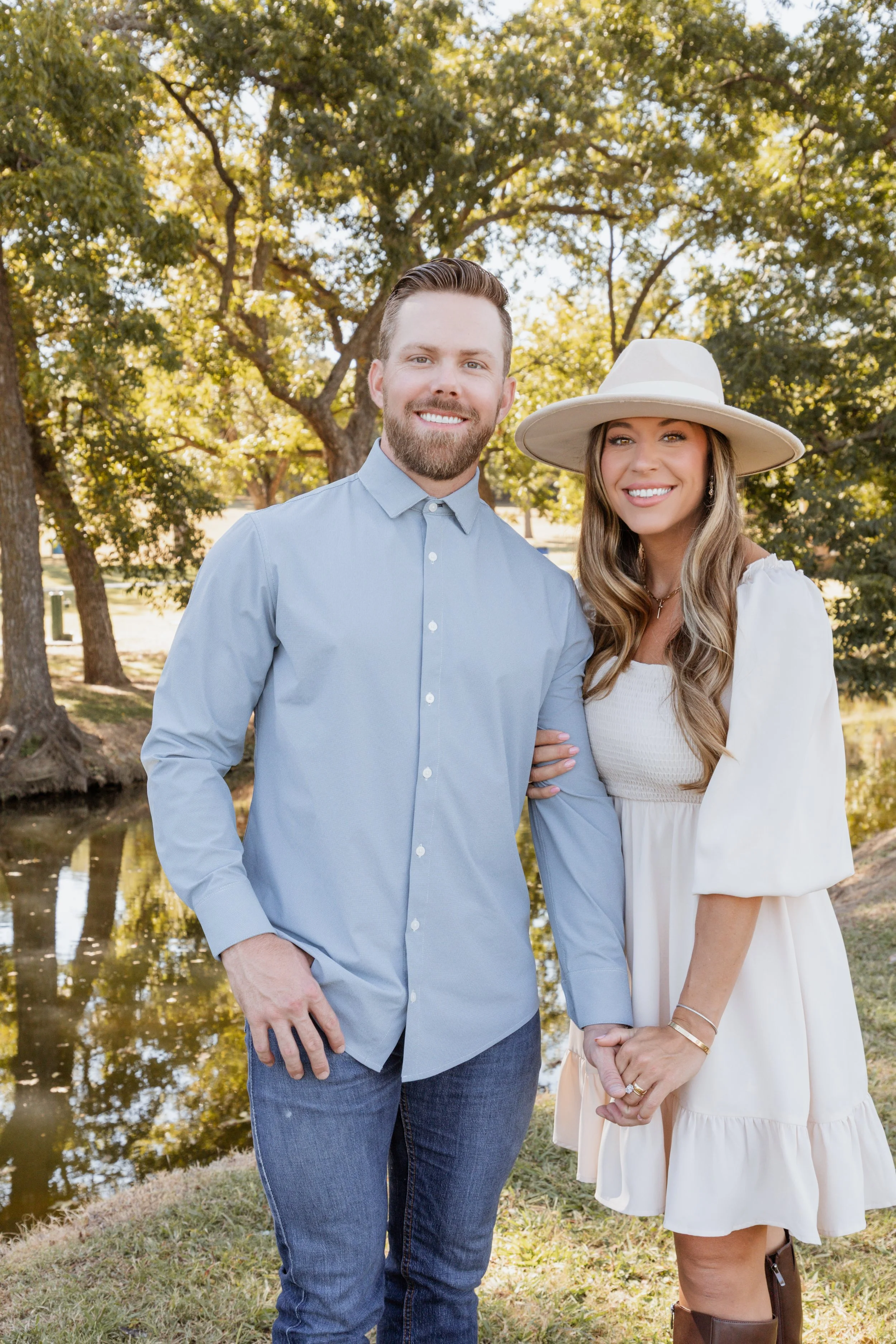 A smiling couple standing outdoors by a tree-lined pond on a sunny day, holding hands, with the woman wearing a large white hat and cream dress, and the man in a light blue shirt and jeans.