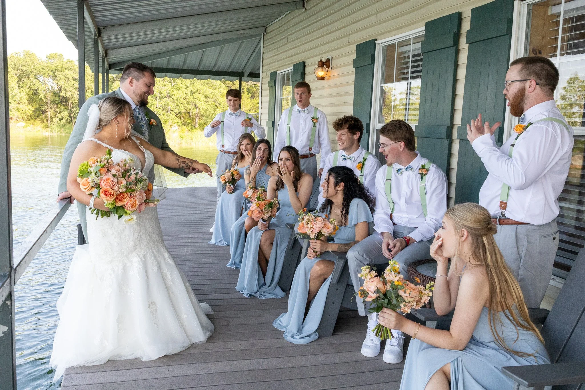 A wedding party on a porch by the water, with the bride and groom in the center, surrounded by bridesmaids and groomsmen. The bride is holding a bouquet, and everyone is wearing pastel-colored attire, with the men in white shirts and suspenders. The 