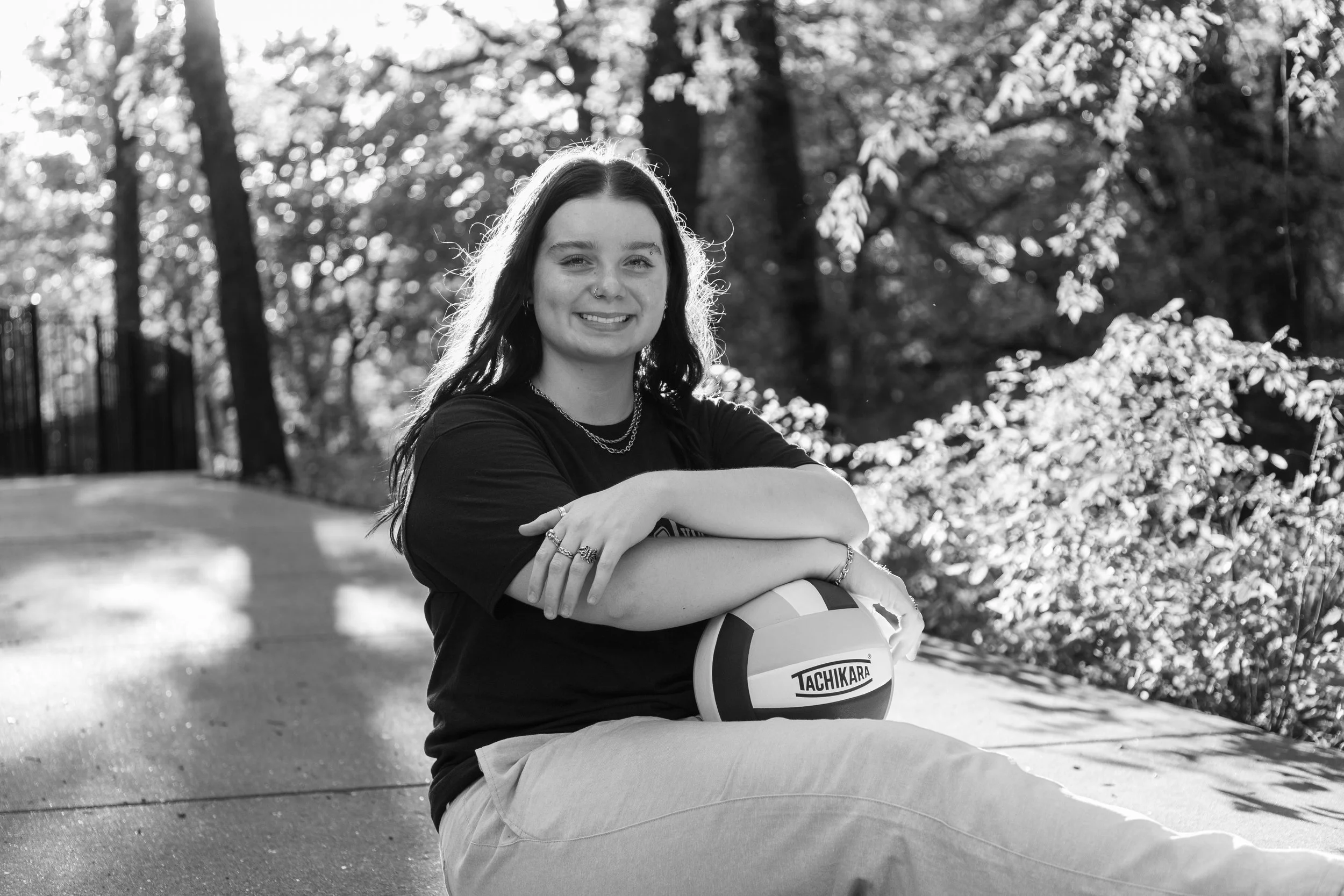 A young woman sitting outdoors on a concrete surface, holding a volleyball, smiling, with sunlight and trees in the background in black and white.