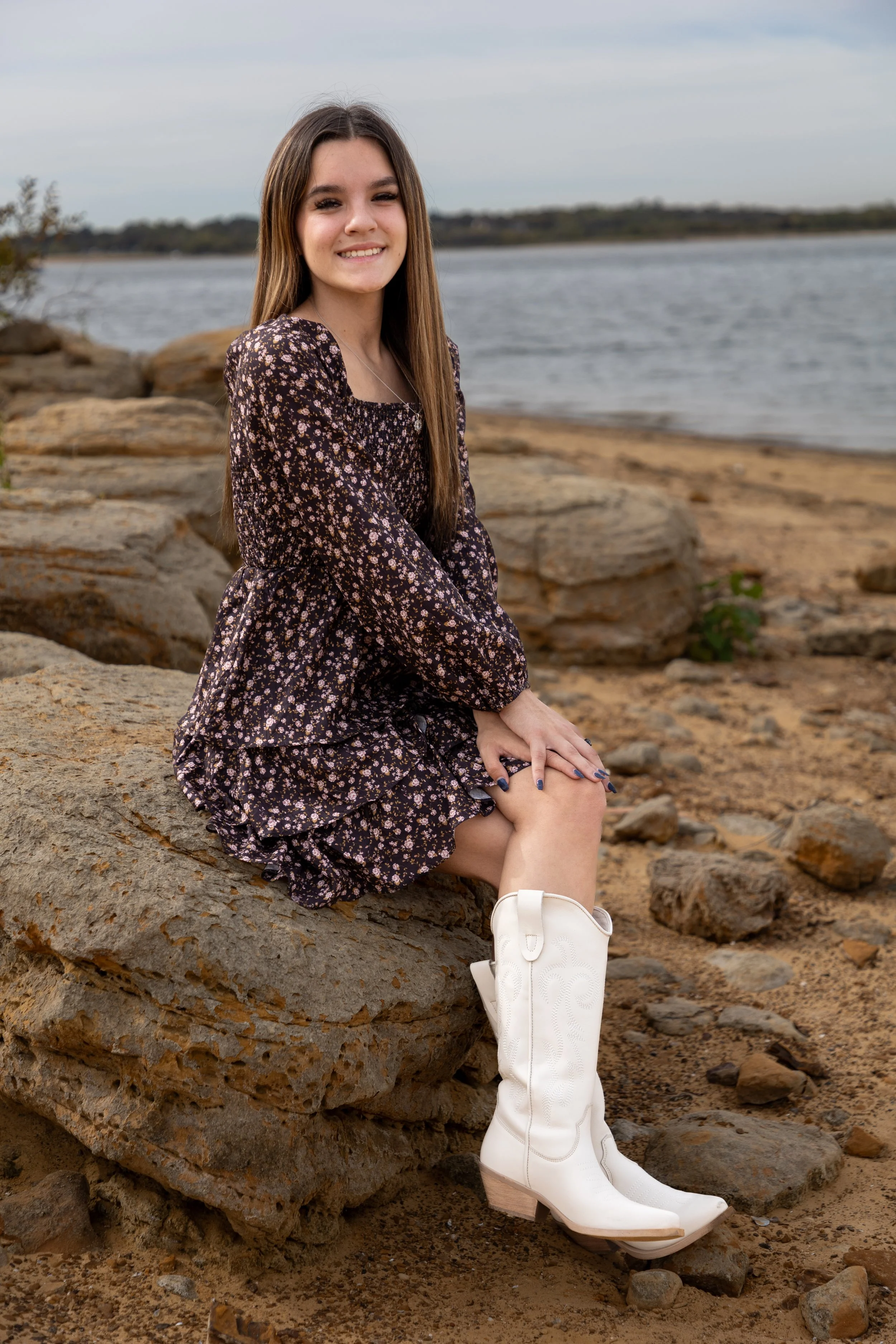 A young woman with long brown hair sitting on a large rock by the water. She is wearing a black floral dress with long sleeves and white cowboy boots, smiling at the camera.