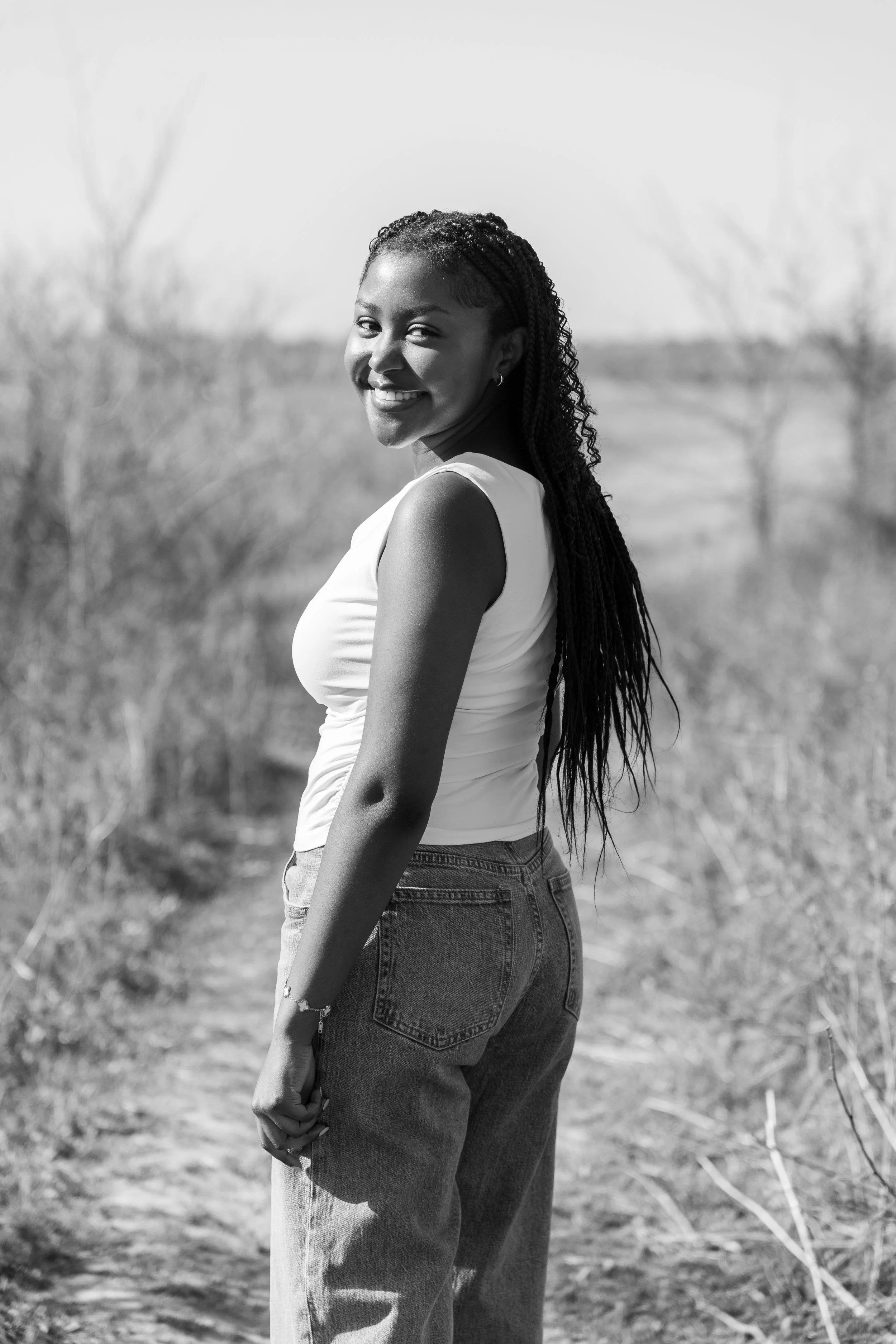 Smiling young woman with long braided hair standing outdoors in a rural setting, wearing a sleeveless top and jeans.