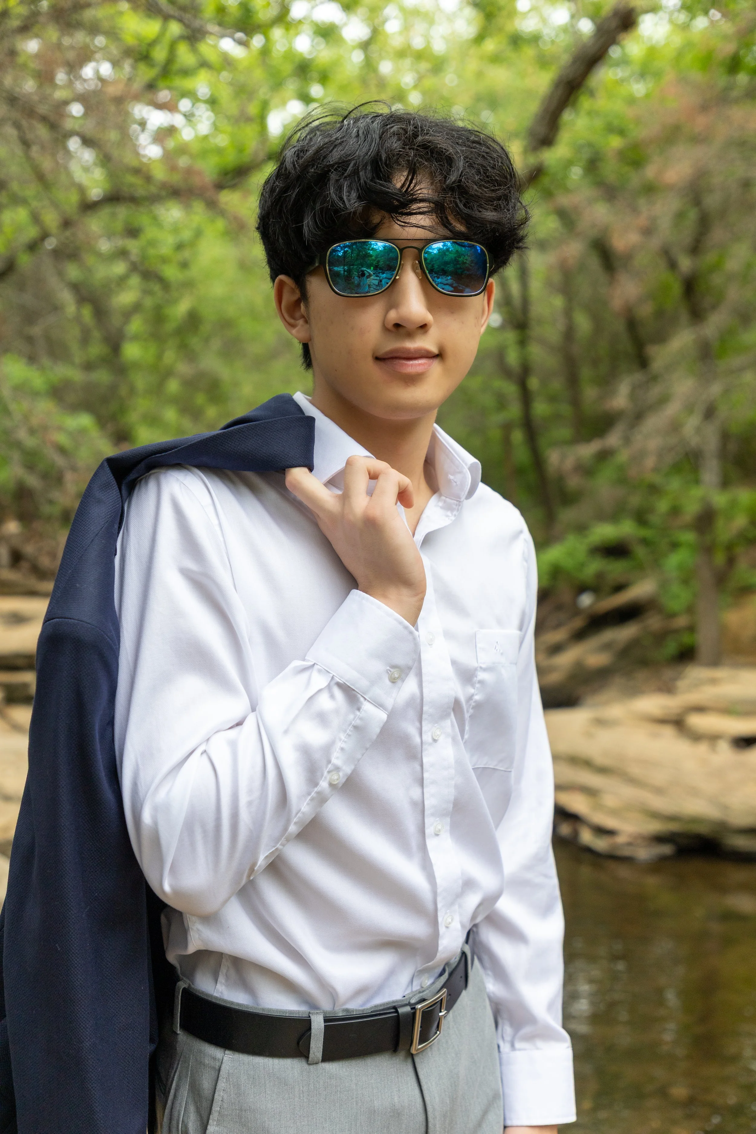 Young man wearing sunglasses, a white dress shirt, gray pants, and a black belt, holding a navy jacket over his shoulder, standing outdoors in a forested area.