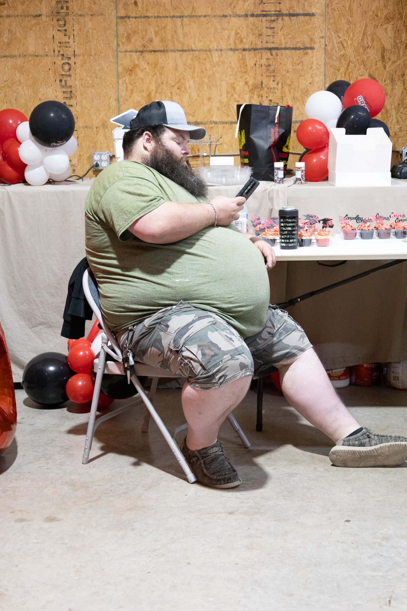 A man with a beard sitting on a folding chair at a table decorated with balloons, holding a smartphone.