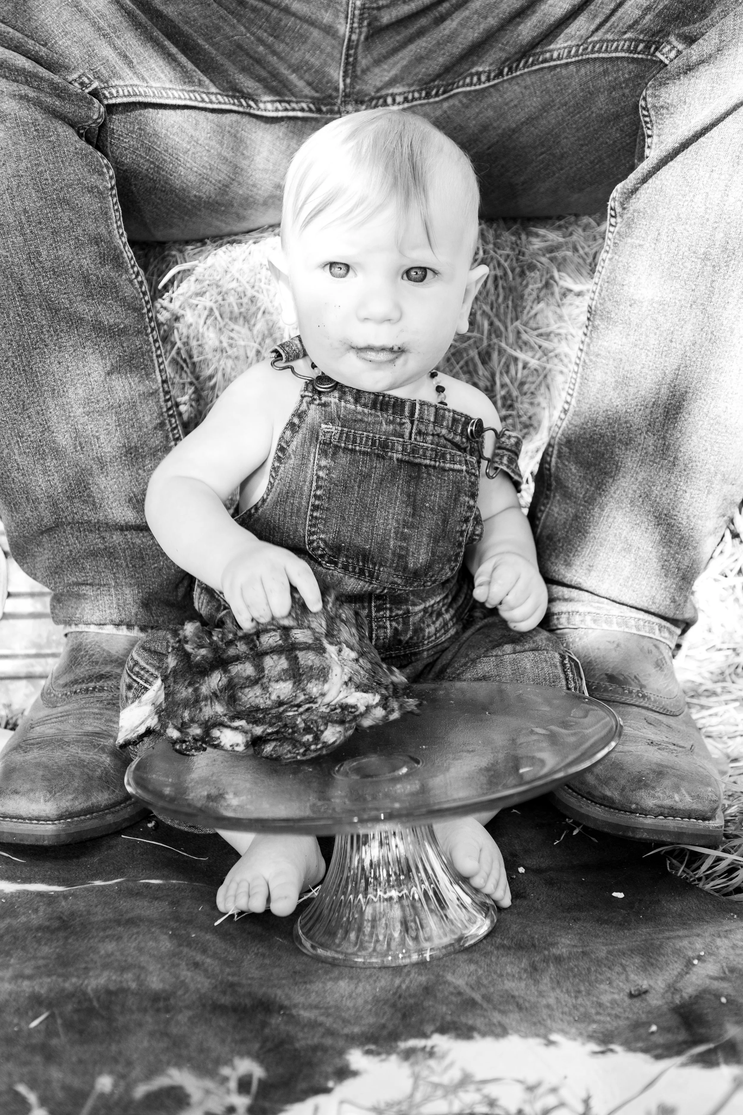 Black and white photo of a baby sitting on a person's lap, wearing overalls and barefoot, holding a piece of food, with a decorative platter in front.