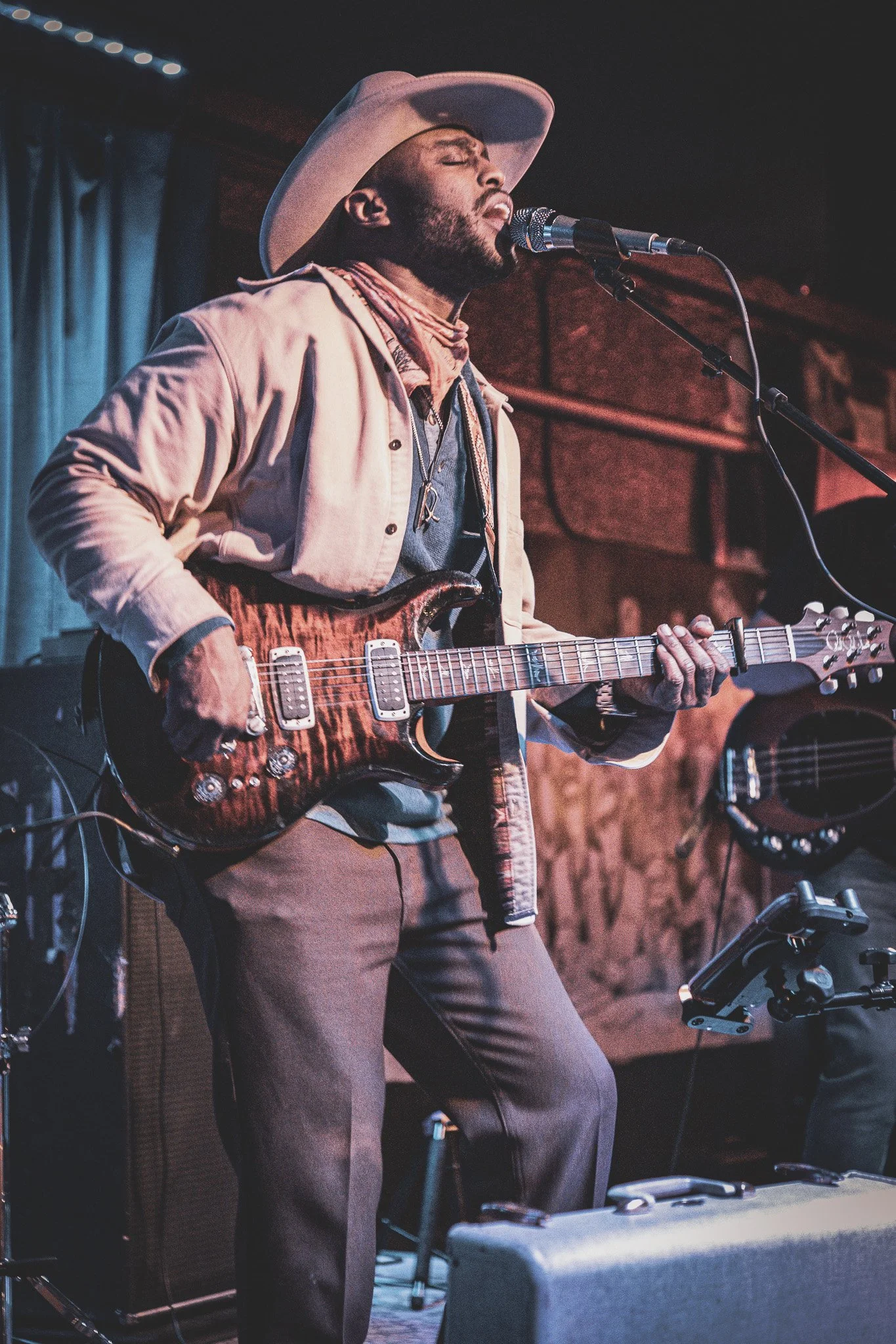 A man wearing a wide-brim hat and a beige jacket plays an electric guitar and sings into a microphone on stage in a music venue.