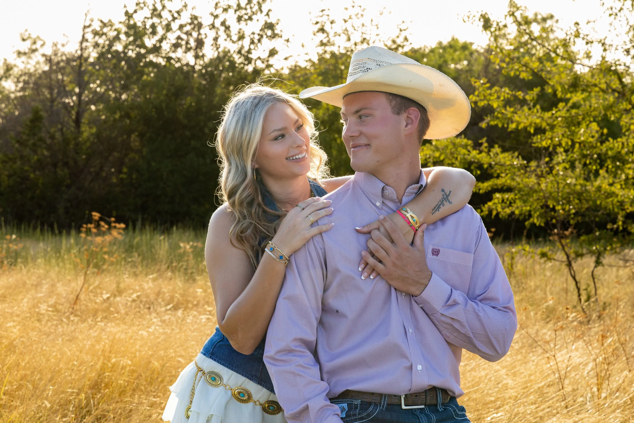 A young woman and a young man are embracing outdoors in a field with trees in the background. The woman has blonde hair and is smiling while looking at the man, who is wearing a cowboy hat and looking back at her.