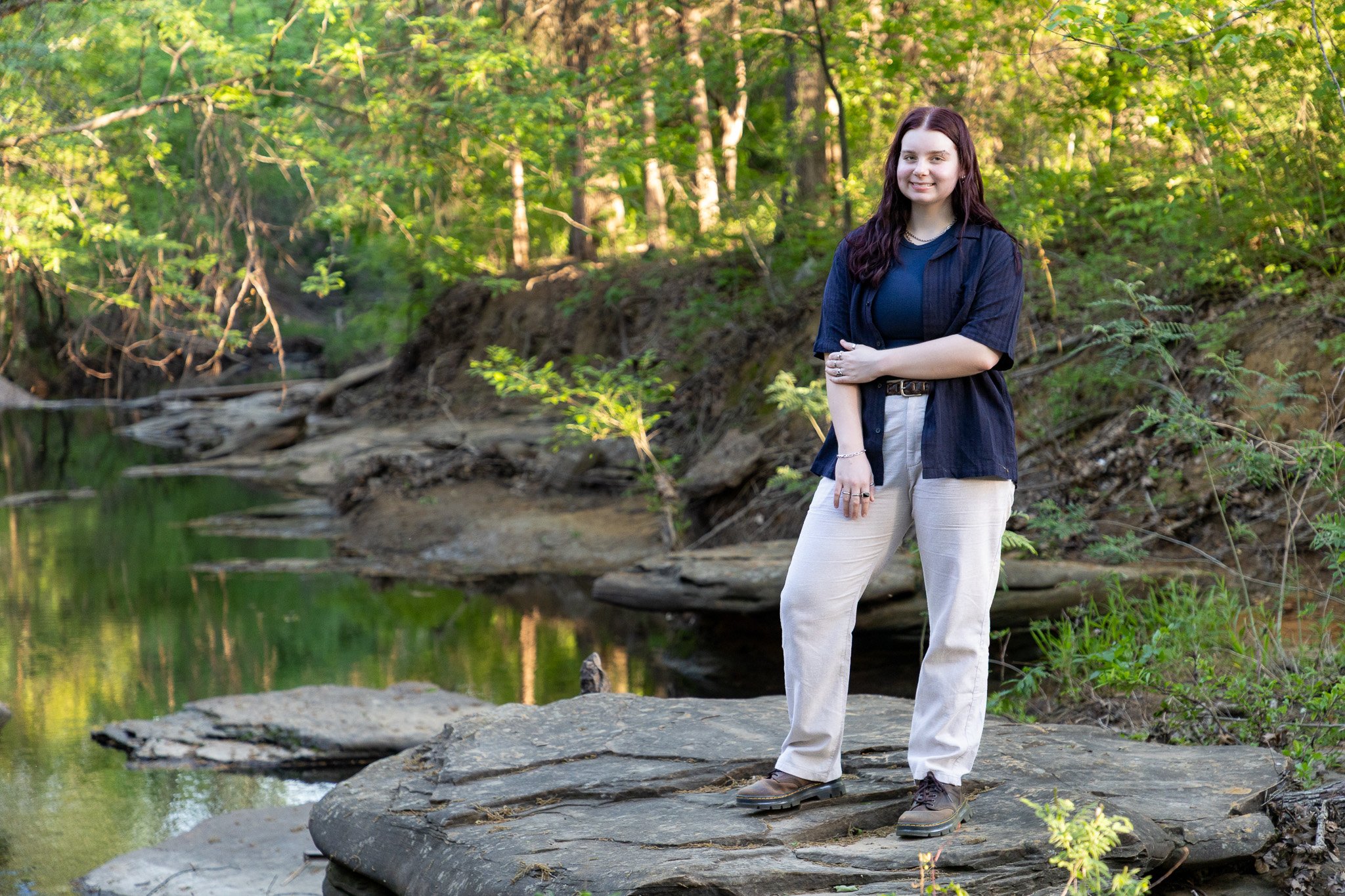 A young woman standing on a large flat rock beside a small stream in a forested area, smiling at the camera, with trees and greenery in the background.