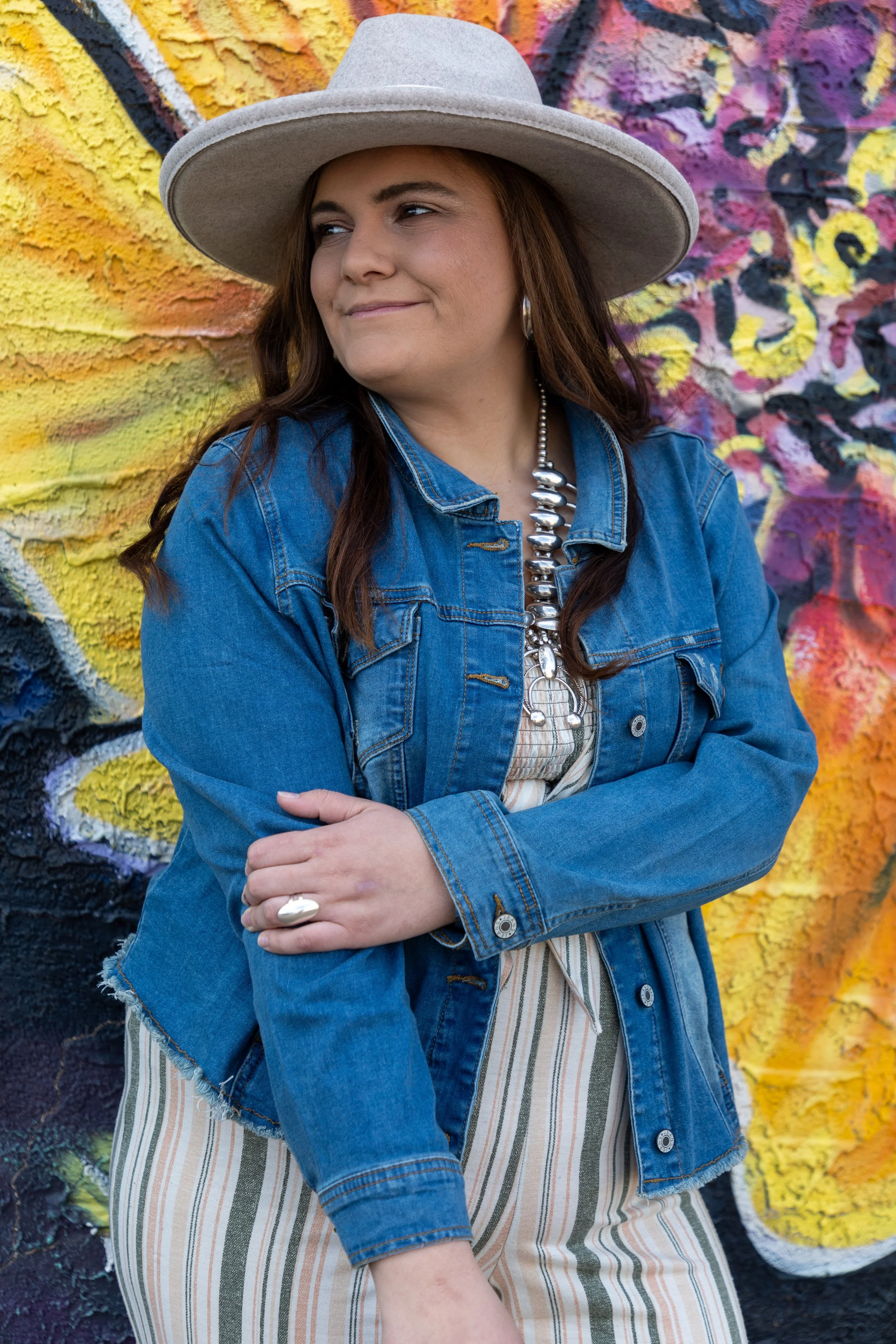 A woman wearing a gray wide-brim hat, a denim jacket, and a chunky metallic necklace, standing in front of a vibrant colorful graffiti wall.