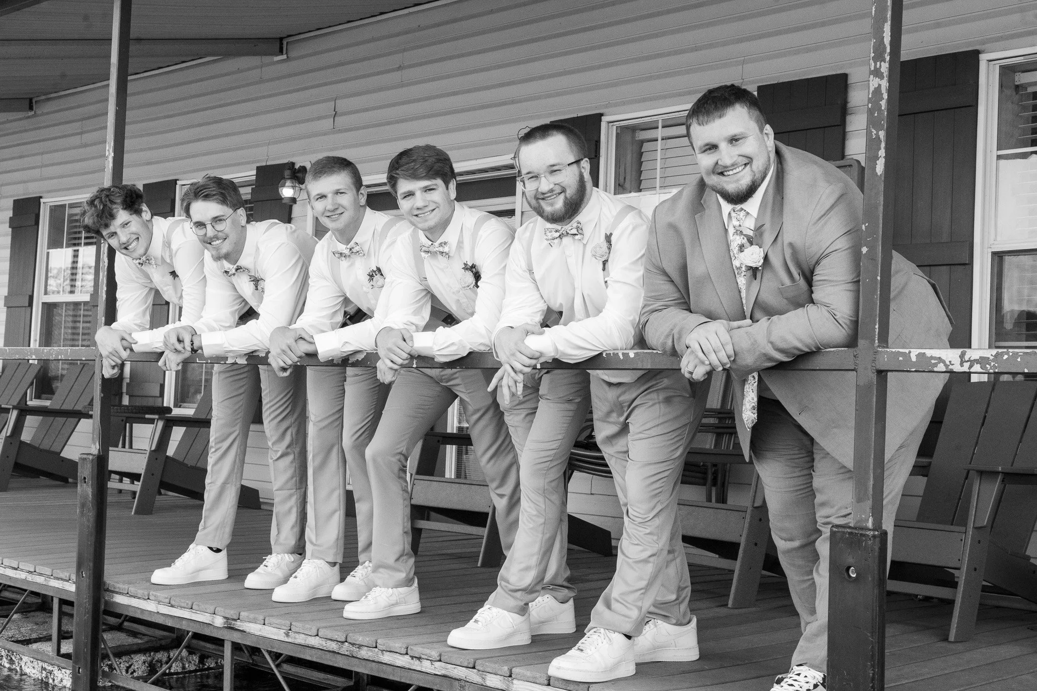 Black and white photo of seven men in formal attire leaning on a railing on a porch, smiling, with a house in the background.