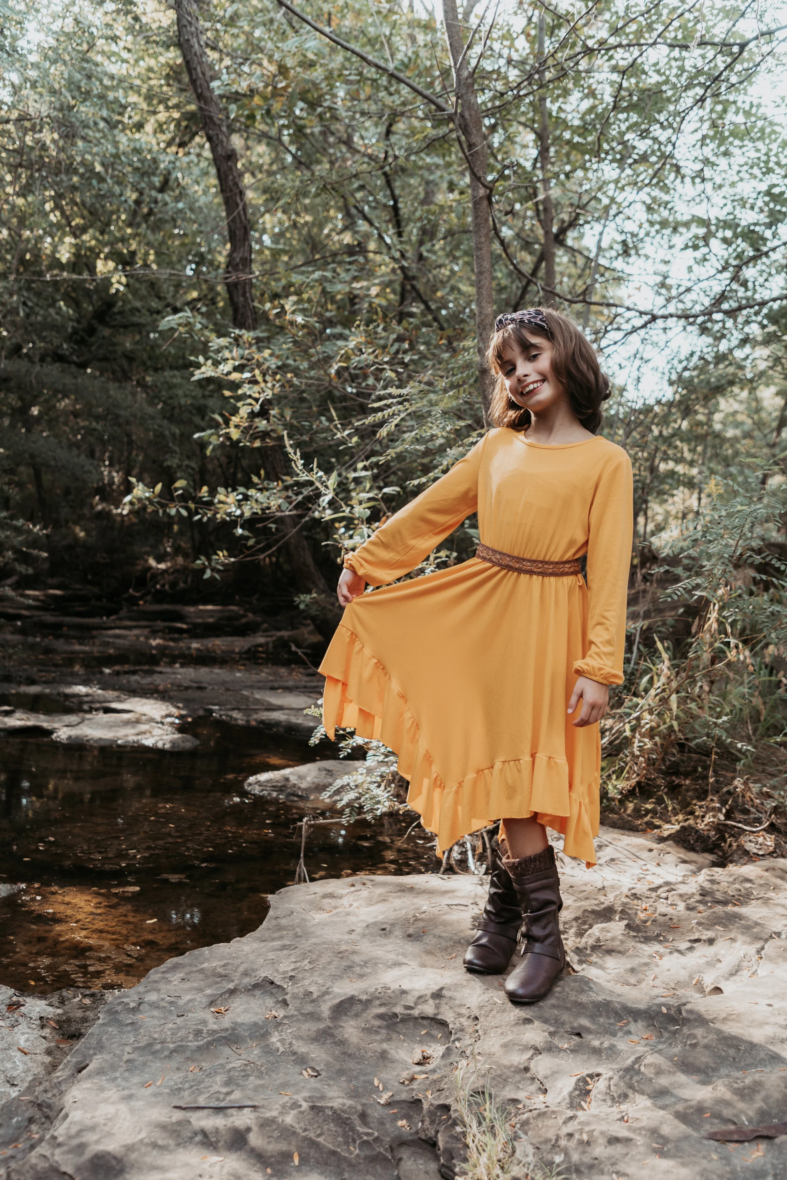 A girl in a yellow dress and brown boots standing on a large rock near a creek in a wooded area, smiling and holding her dress.