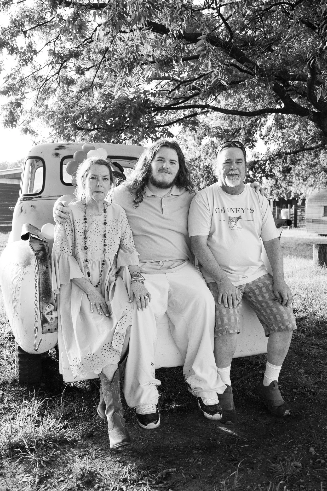 Black and white photo of three people sitting on the back of a pickup truck outdoors under a tree. The woman on the left wears earrings, a long dress, and cowboy boots. The young man in the middle has long hair and a beard, wearing a polo shirt and p