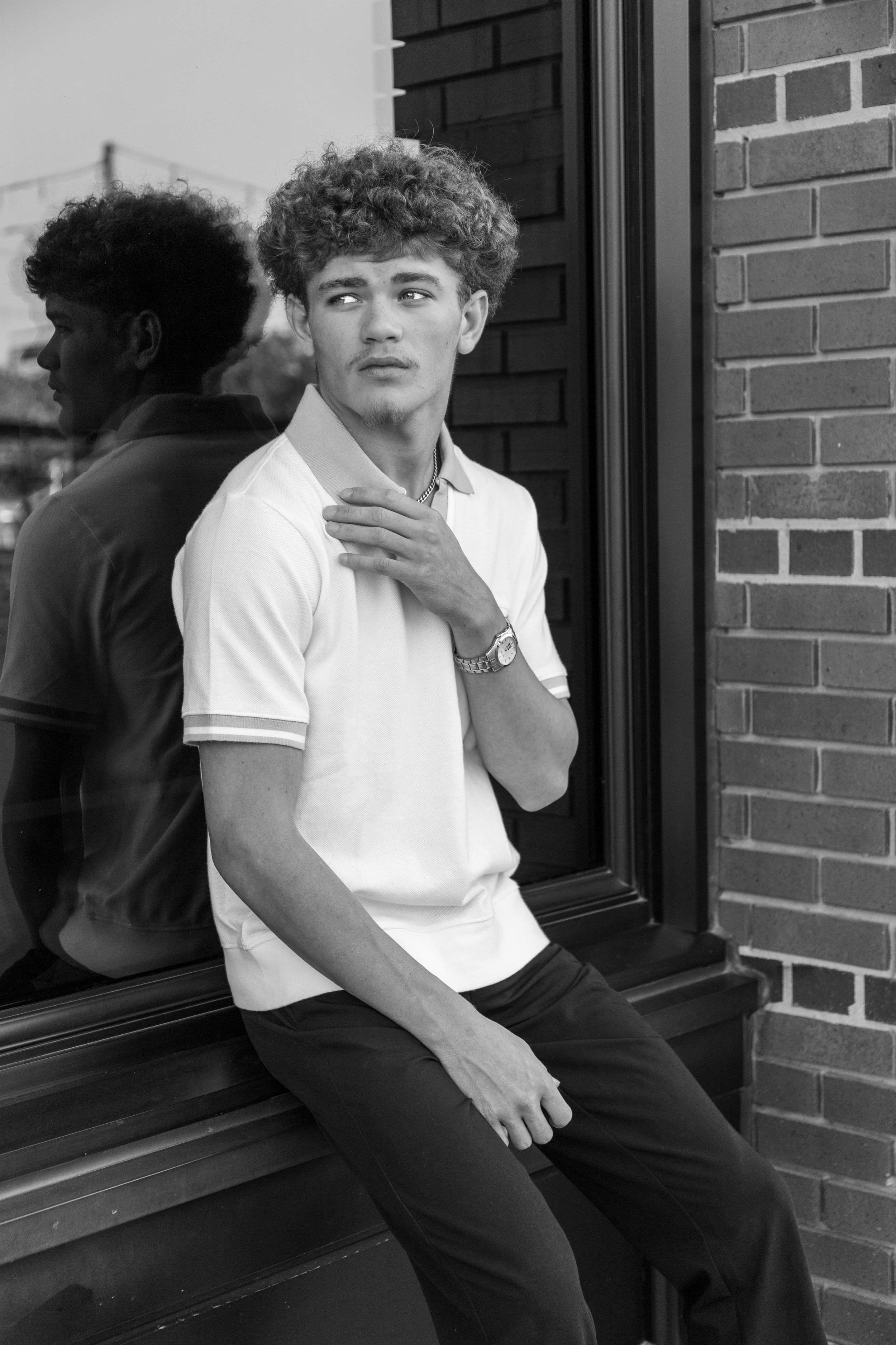 A young man with curly hair, wearing a white polo shirt, sitting on a ledge beside a large window, looking off to the side with one hand on his shoulder, in a black and white photo.