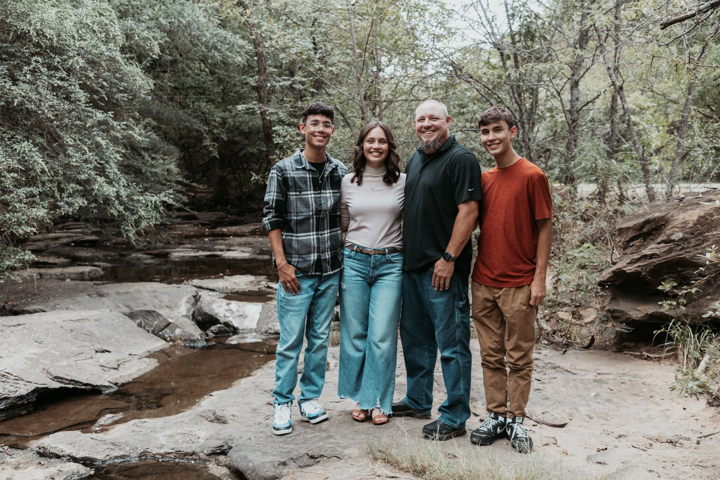 A family of five standing on rocks by a creek in a forested area, smiling at the camera.