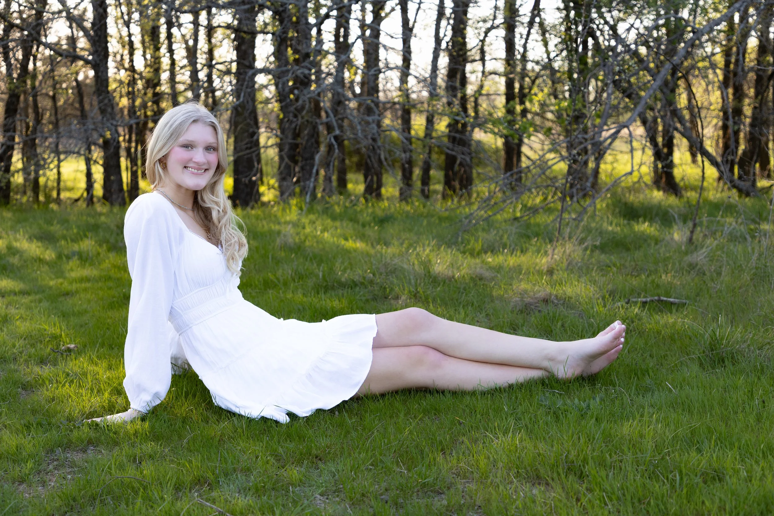 A young woman in a white dress sitting on green grass in a forested area, smiling at the camera.