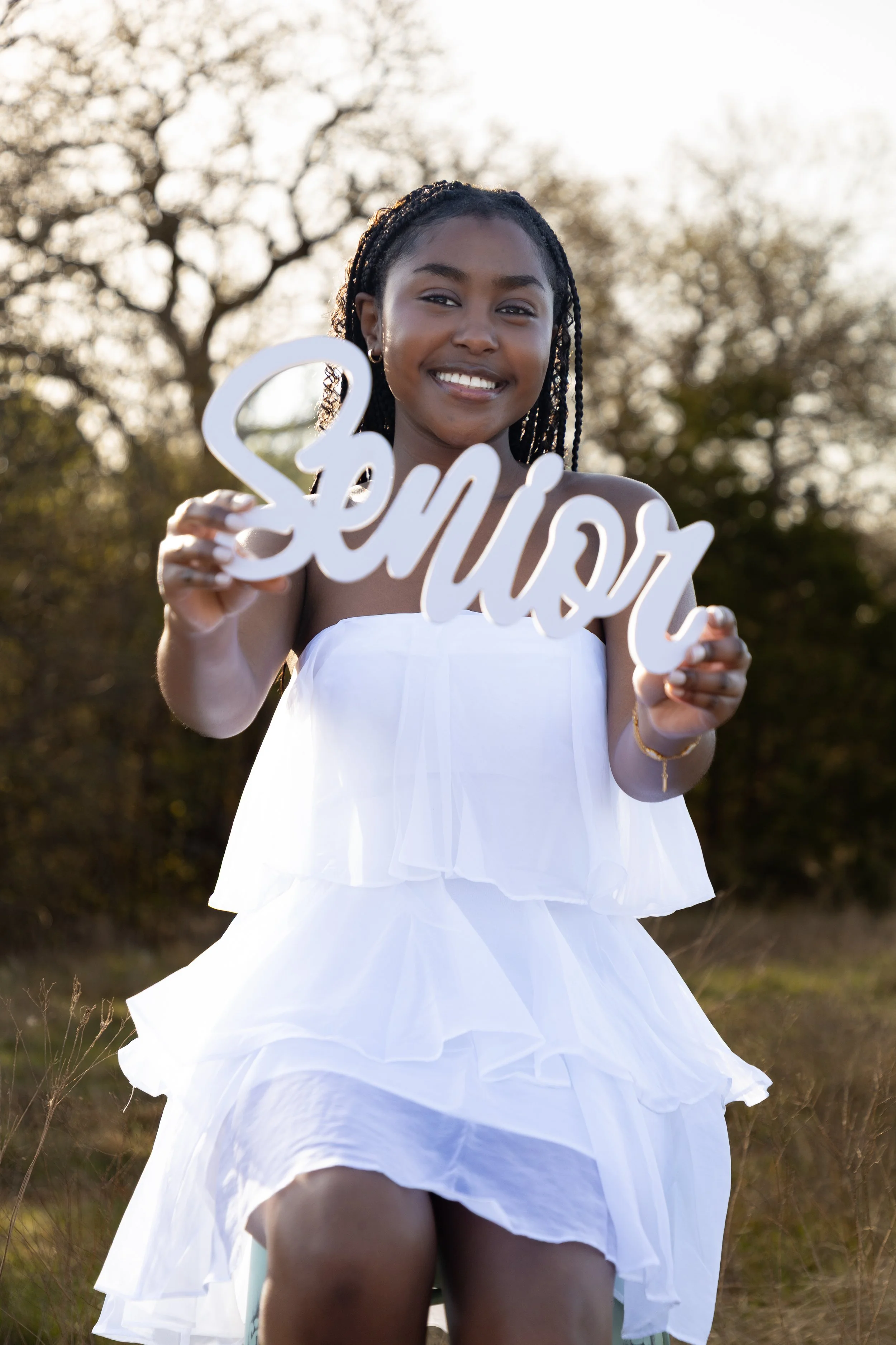 A young girl in a white dress sitting outdoors, smiling, and holding a sign that says 'senior' during the daytime.