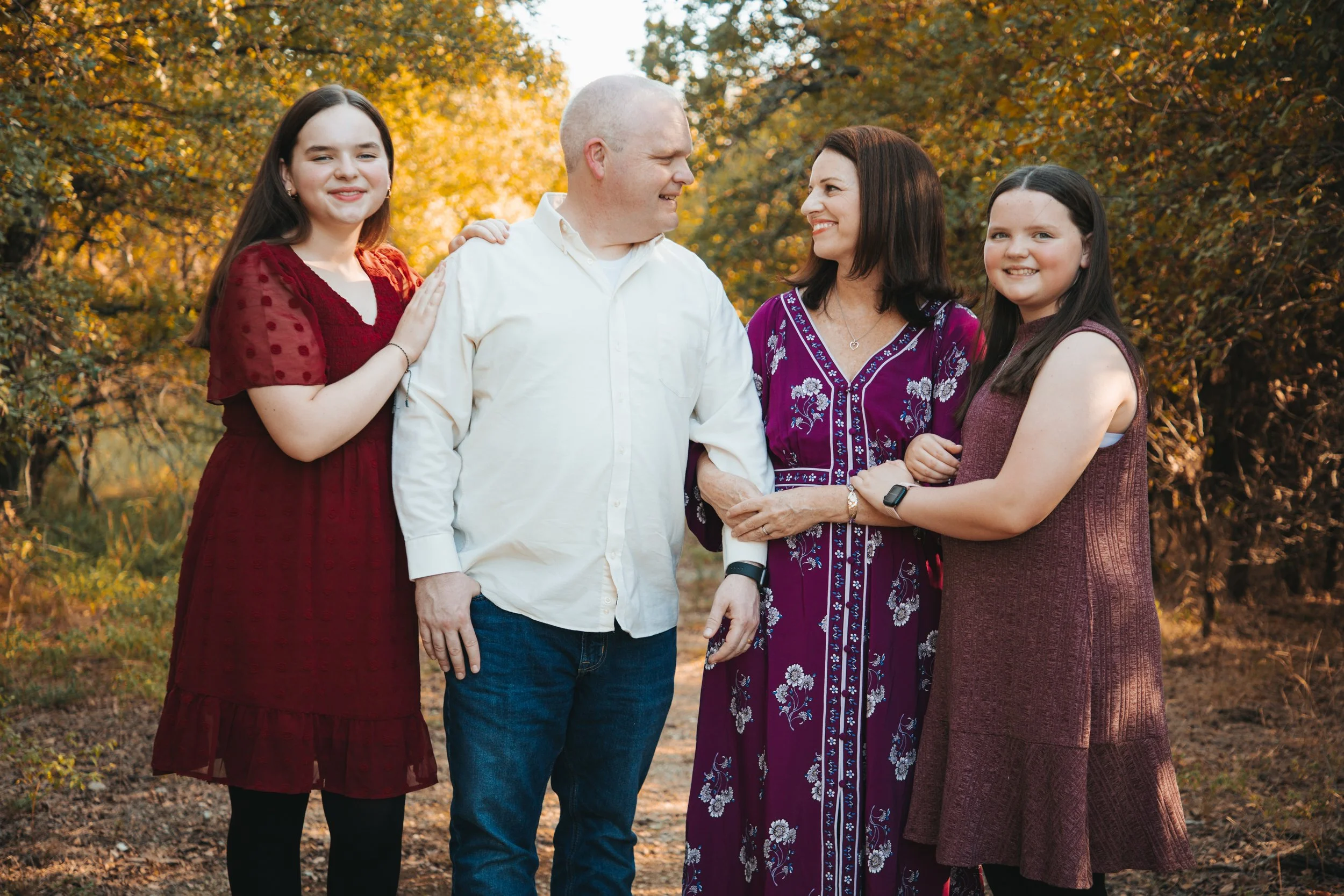 A family of five standing outdoors during fall, with trees and golden leaves in the background. The family members are smiling and interacting, with the two young women on each side, a man in the middle, and a woman next to him.