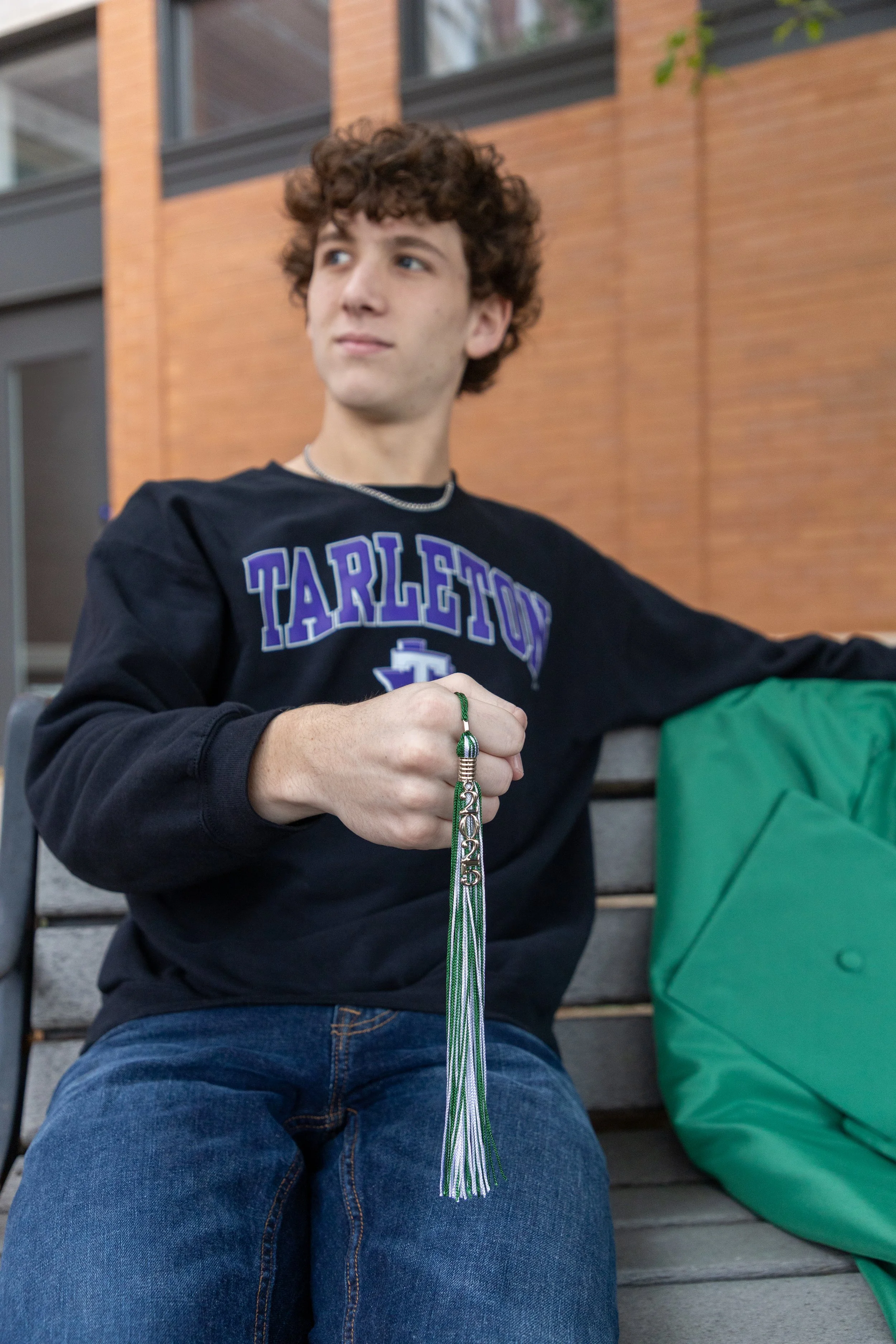 A young man with curly brown hair sitting on a park bench holding green and white tassels. He is wearing a black sweatshirt with purple text 'TARLETOI' and blue jeans, with a green bag beside him.