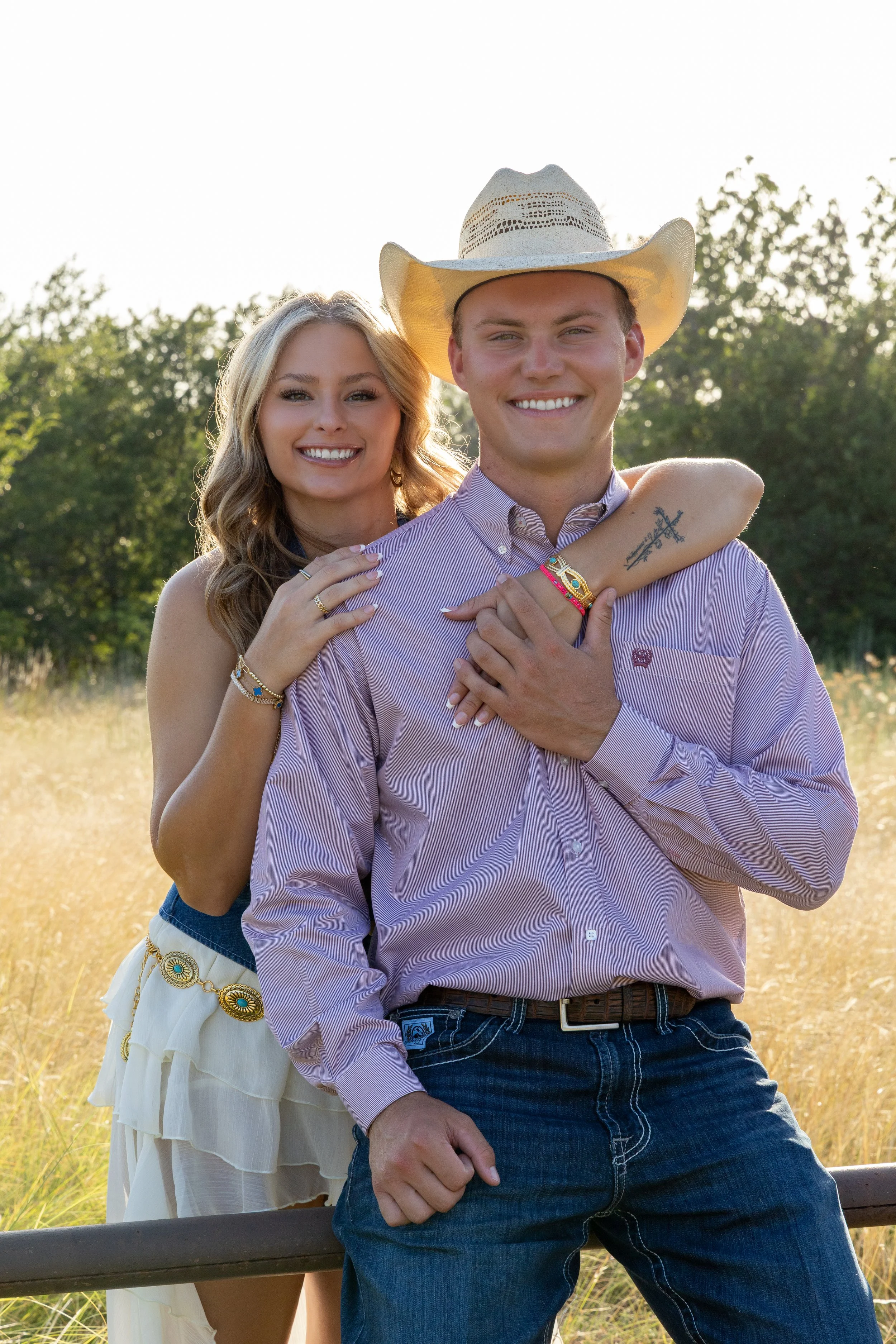 A young couple outdoors in a field, smiling, with the woman standing behind the man with her arm around his shoulder. The man wears a cowboy hat, a light striped shirt, and jeans. The woman has long blonde hair, wears a sleeveless top and a layered w