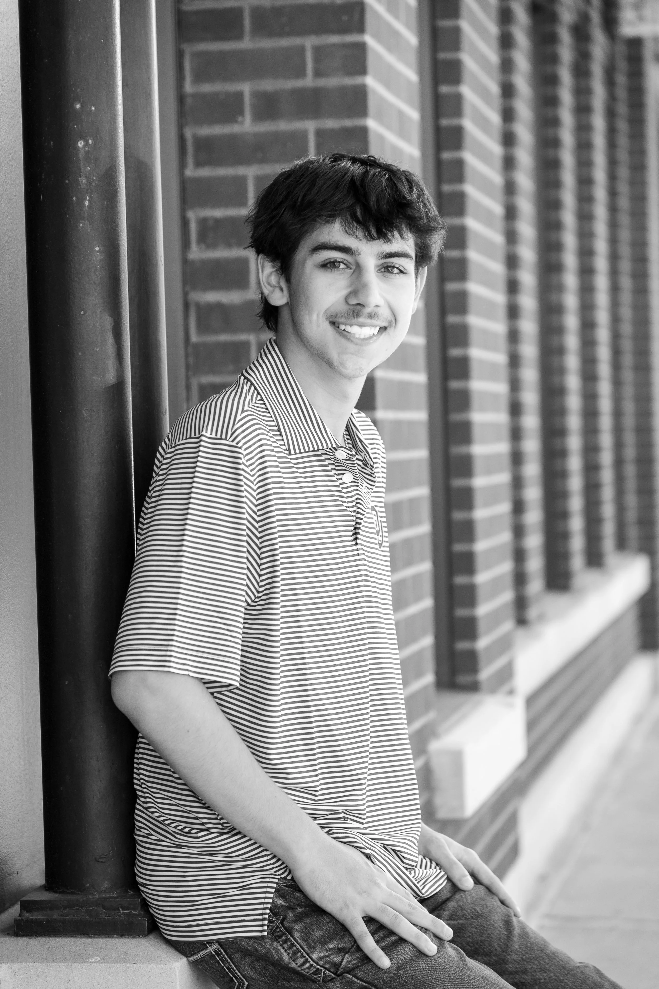 A young man with dark hair and a striped polo shirt leaning against a brick wall and smiling.